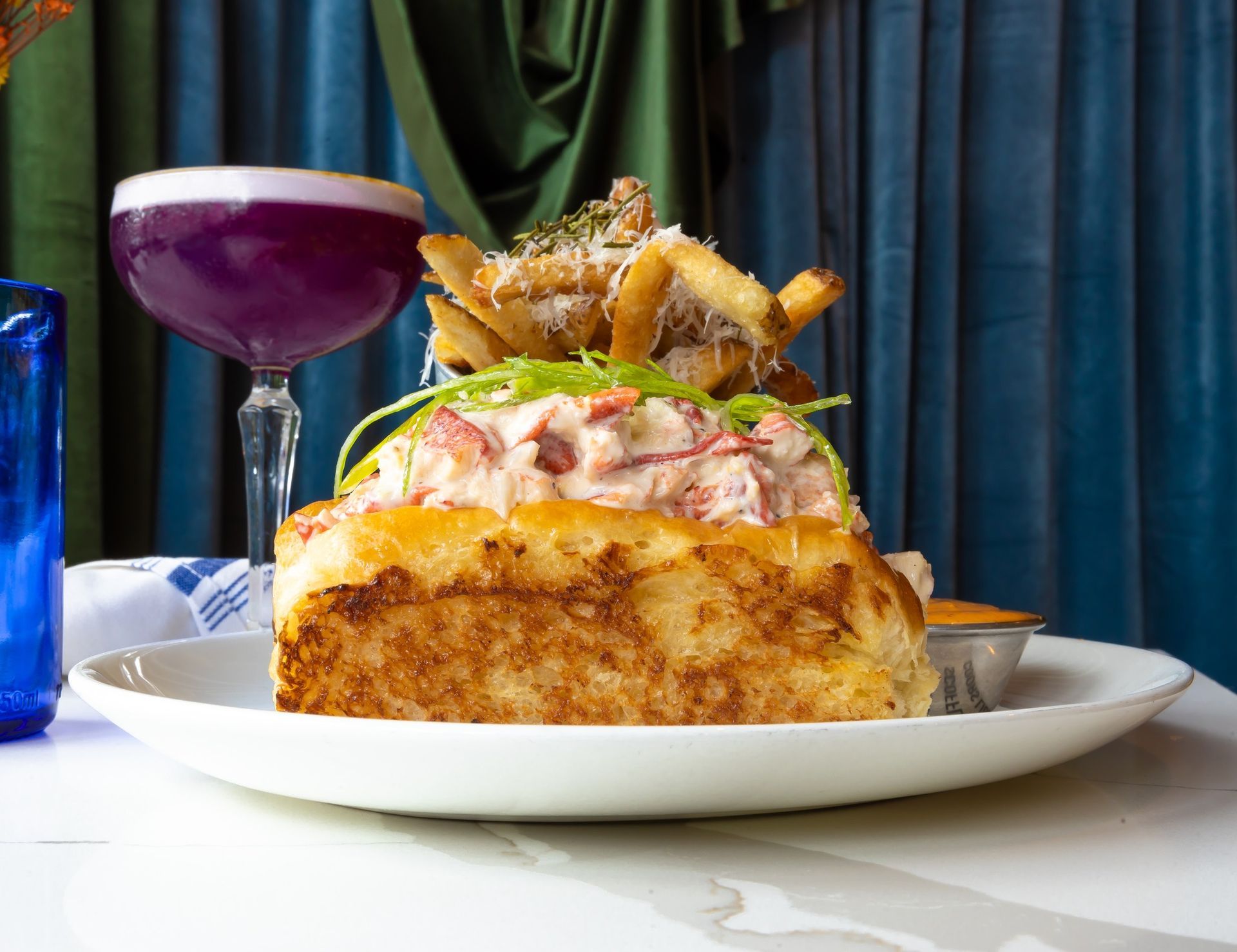 Lobster roll sandwich with fries and purple cocktail on a plate.