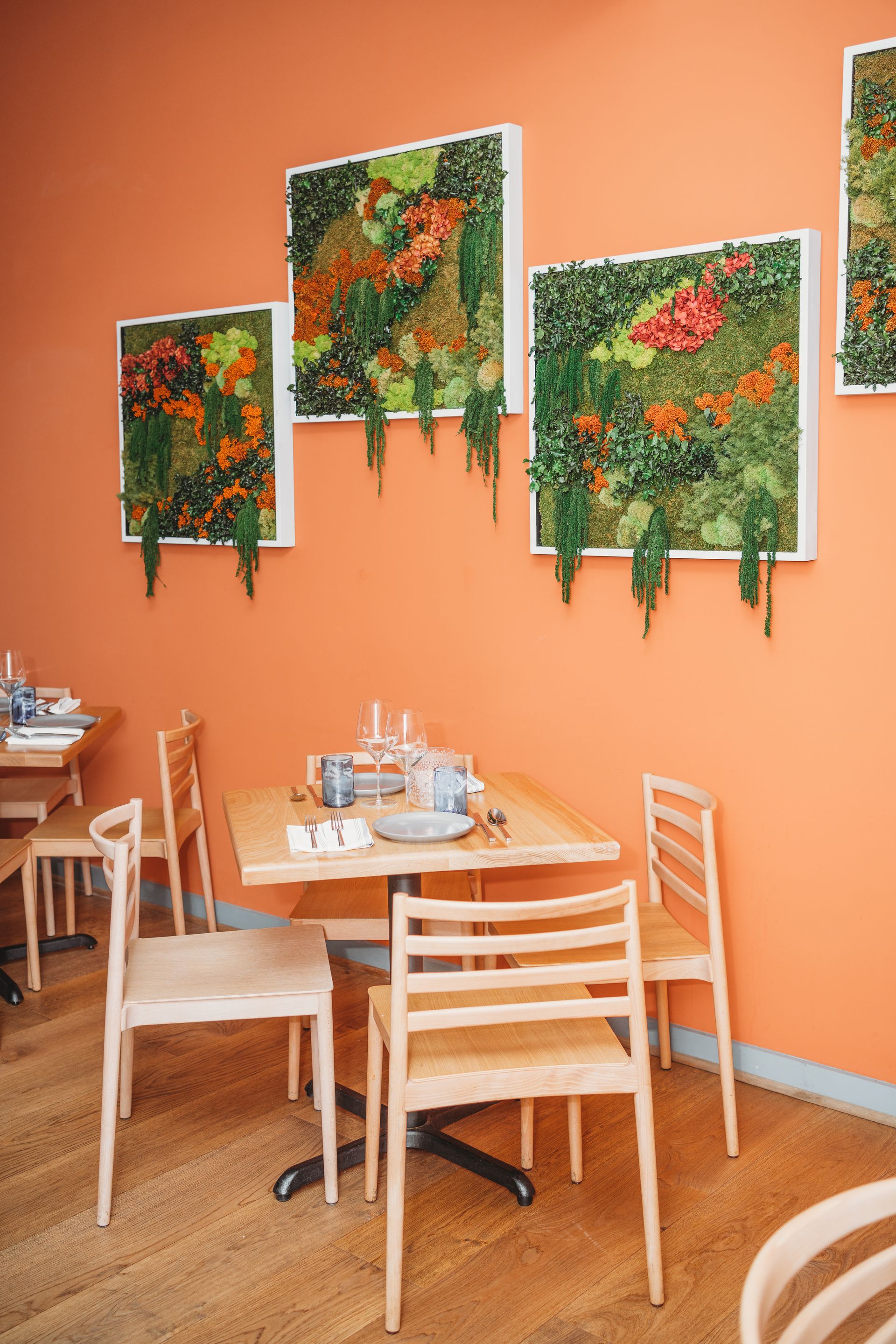 Restaurant dining area with orange walls, wooden tables and chairs, and green plant art.