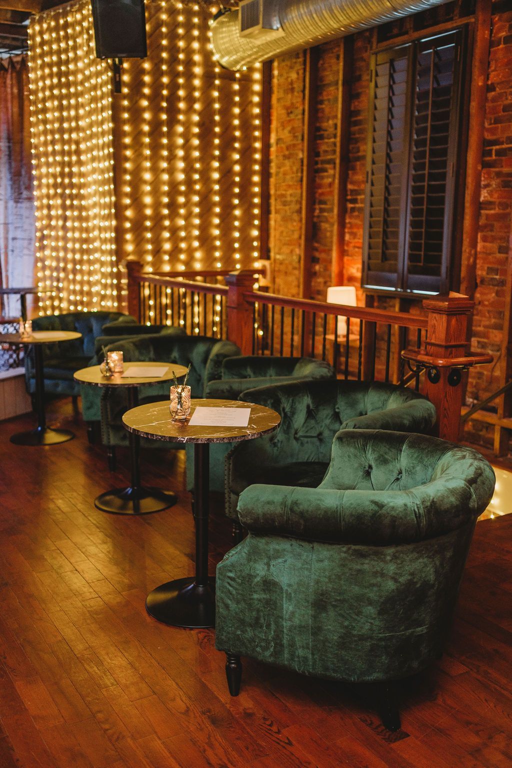 Cozy lounge area with green velvet chairs, small tables, brick wall, and string lights.