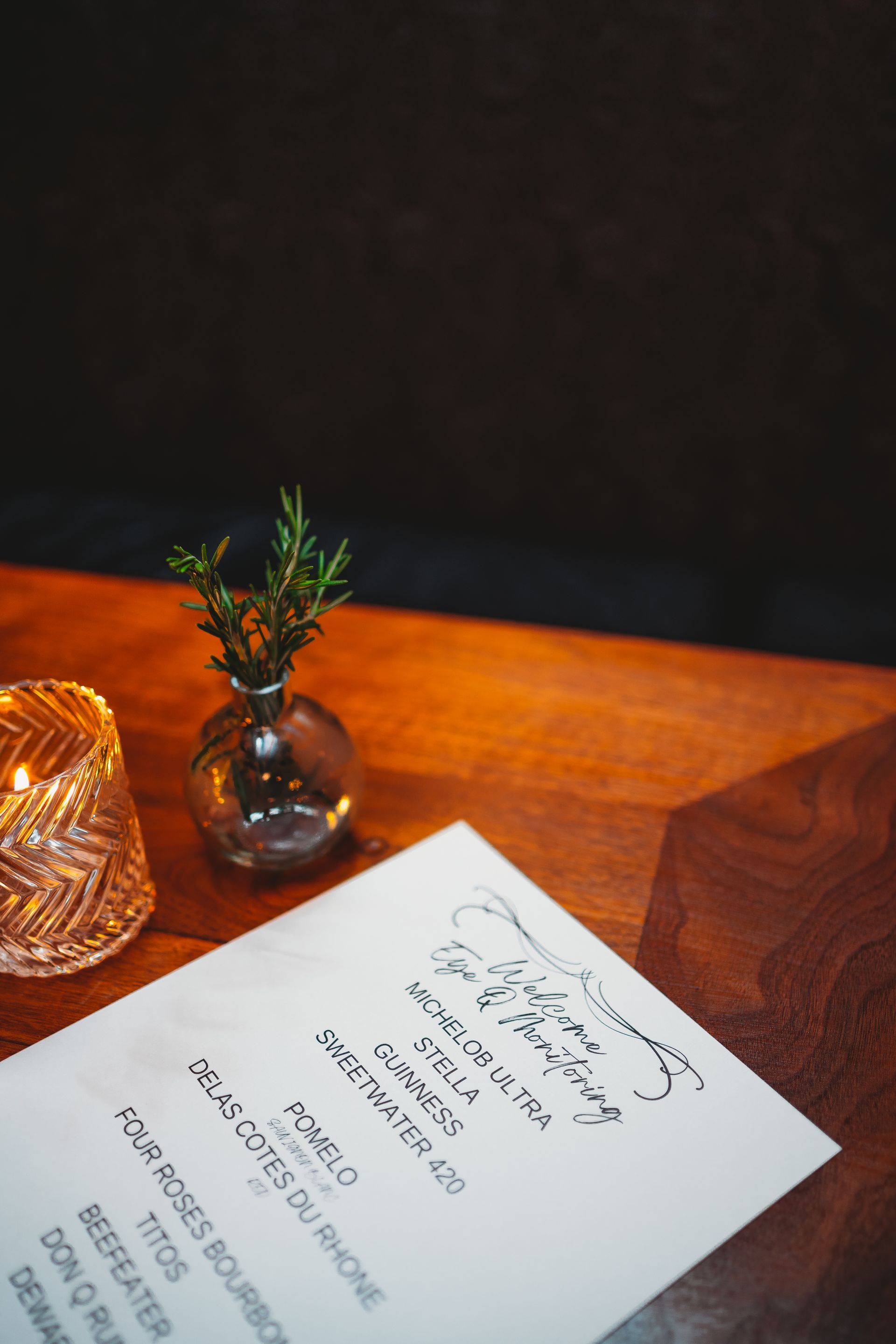 A menu on a wooden table, with a small vase of greenery and a lit candle.