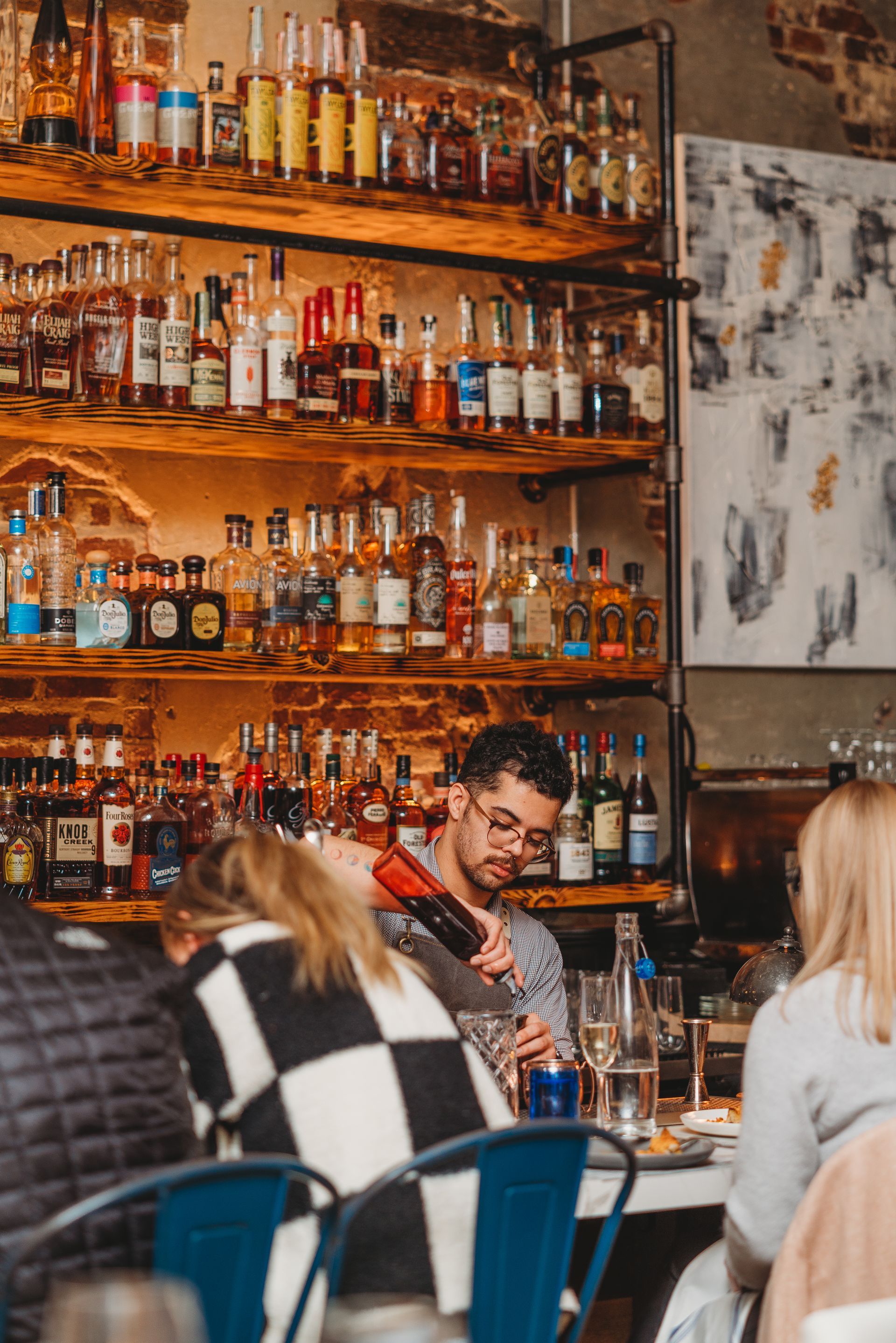 Bartender preparing a drink at a bar with patrons. Shelves stocked with liquor. Rustic decor.