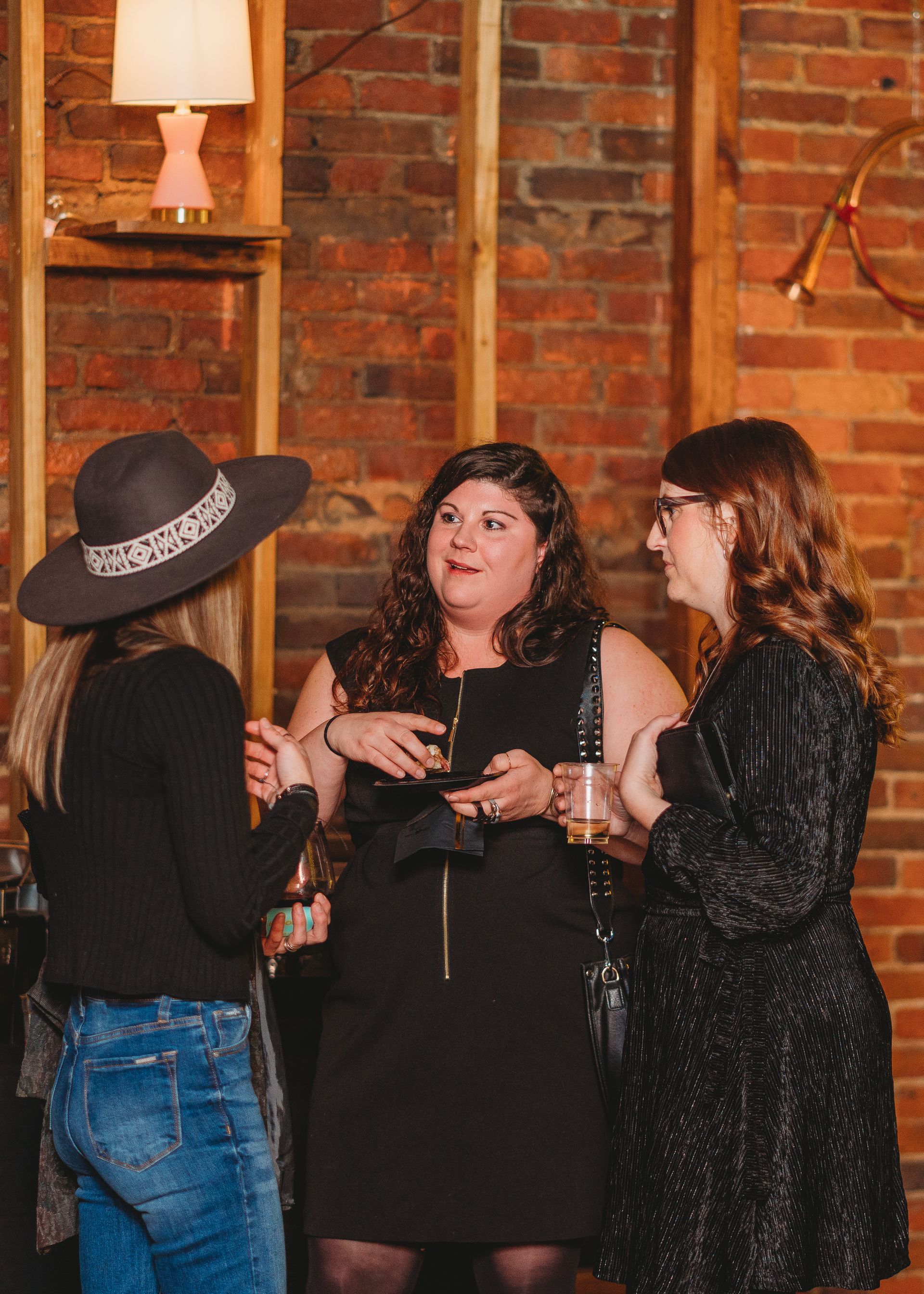 Three women talking at an event, standing in front of a brick wall. One wears a hat, the other two have drinks.