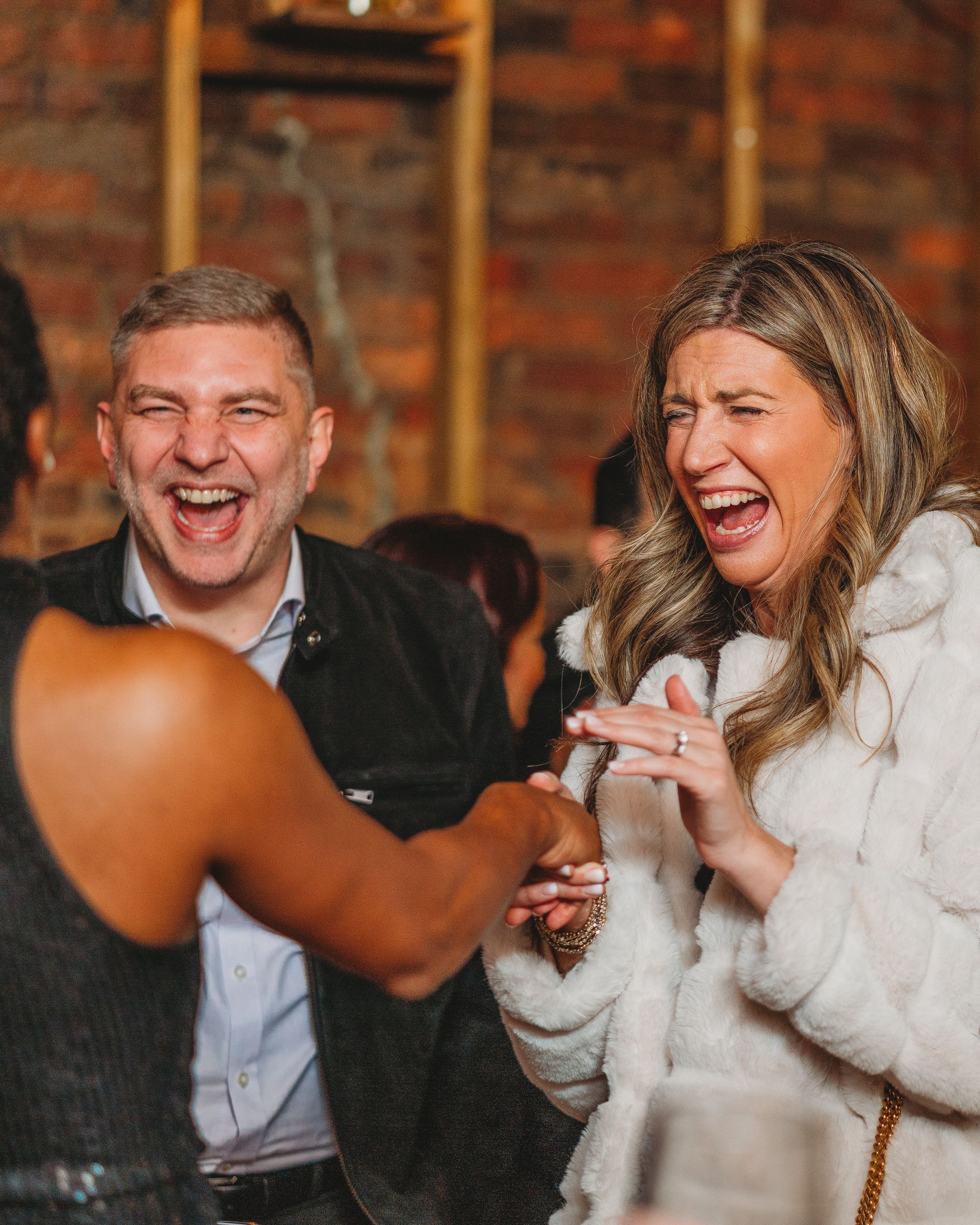 People laughing at an event. A woman in a white coat and a man in a black jacket and a light blue shirt are laughing. A person in a black outfit is next to them.