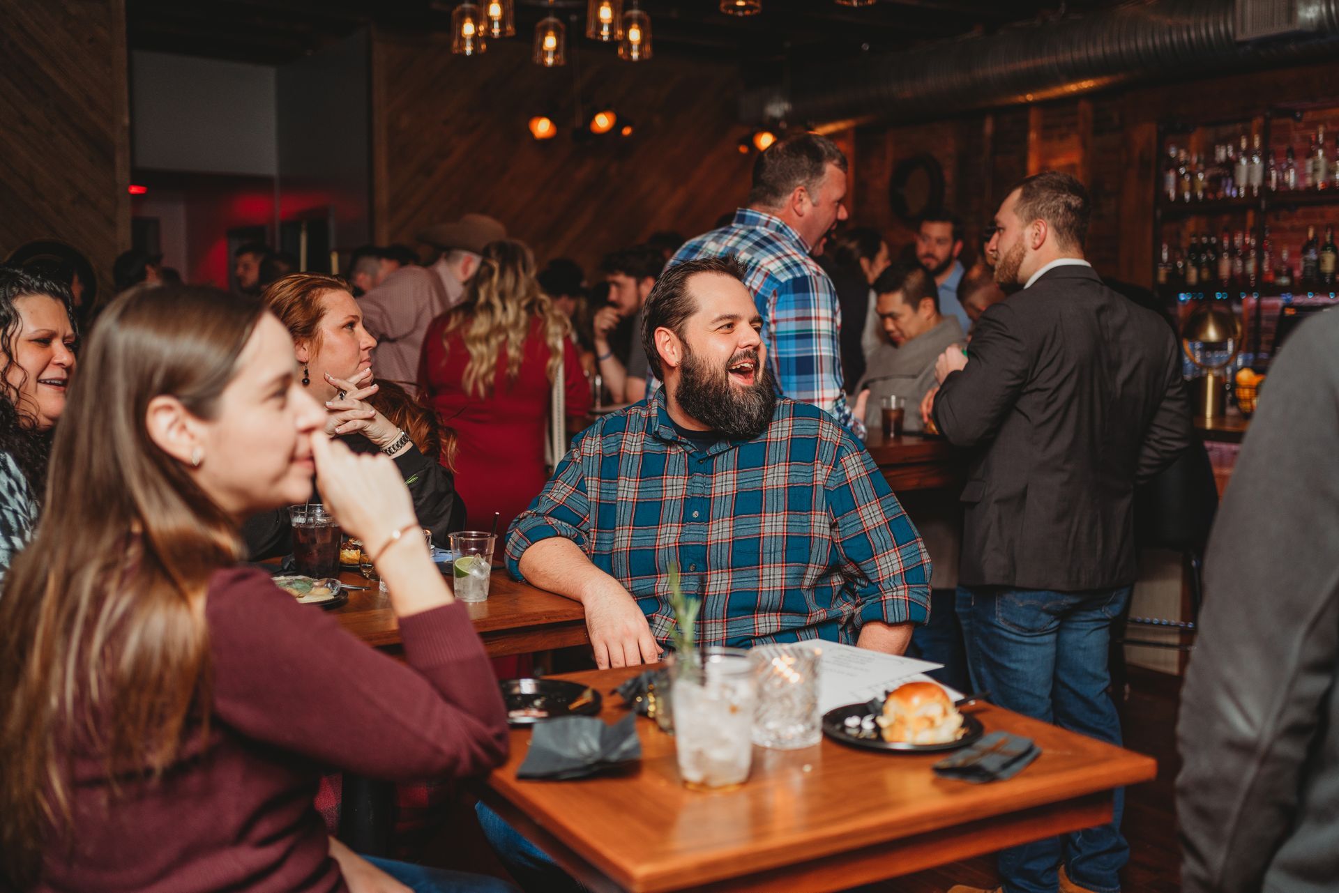 People at a bar, laughing and socializing. Wooden tables, dim lighting, drinks visible.