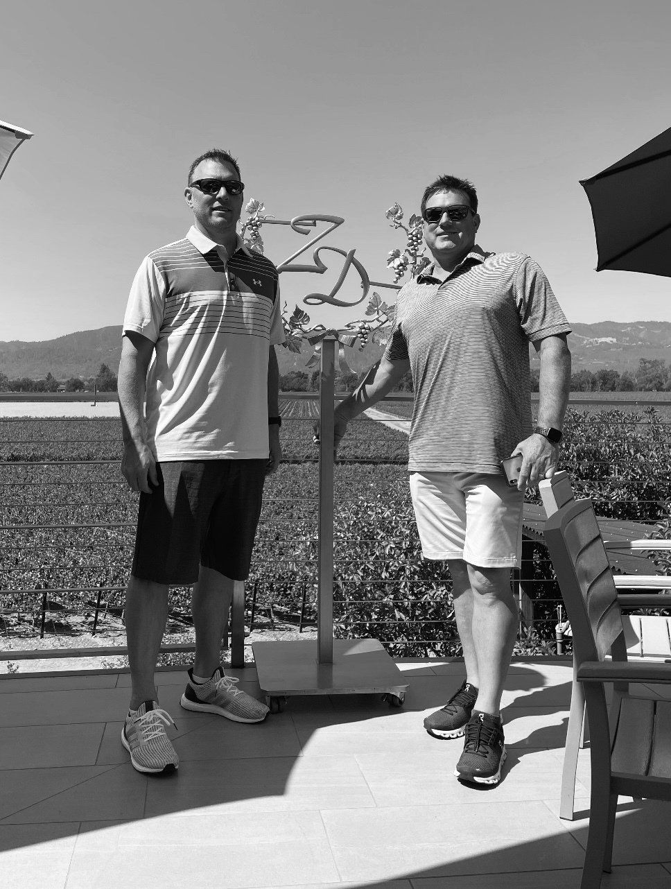 Two men stand near a vineyard sign. One wears shorts, the other white shorts. Sunny day.