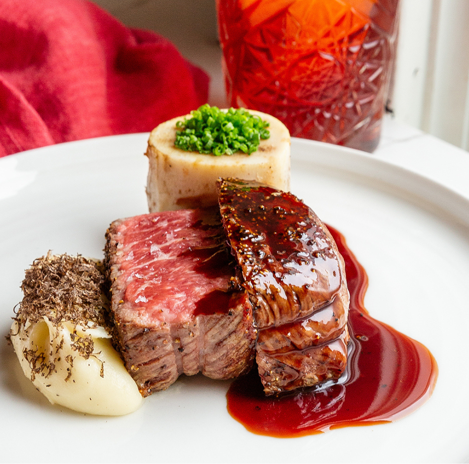 Plate of steak with mashed potatoes, sauce, and a drink in the background.