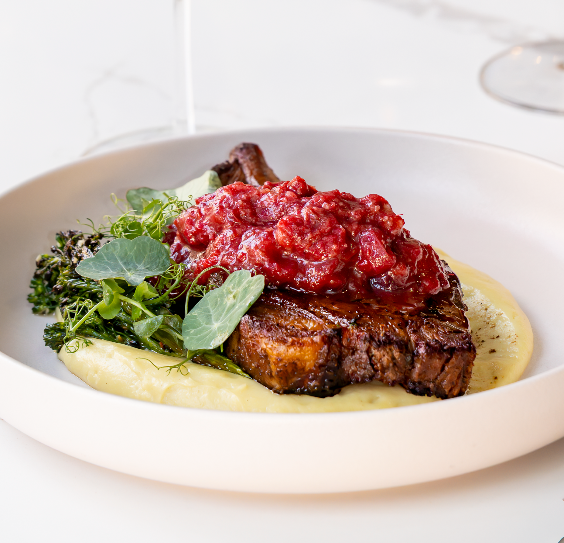 Seared steak topped with red sauce, served with mashed potatoes, greens, and in a white bowl.