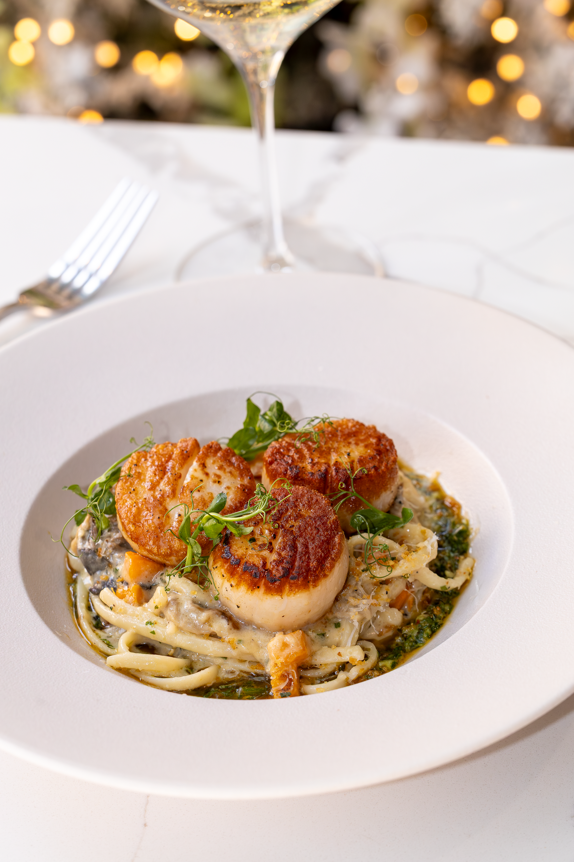 Seared scallops over pasta with pesto, served in a white bowl with a glass of white wine and a fork.
