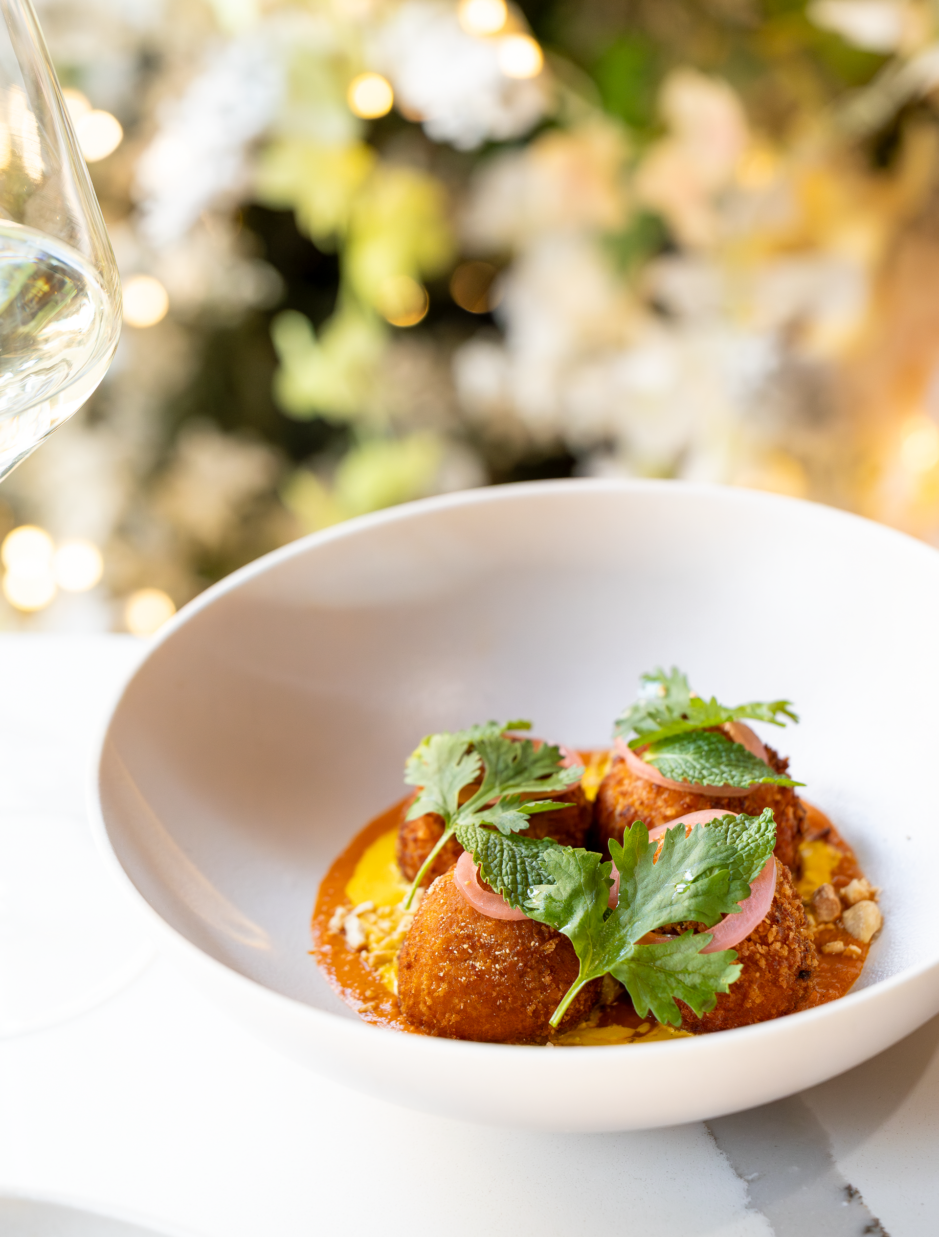 White bowl of food, topped with herbs, with wine glass. Yellow and white blurred background.