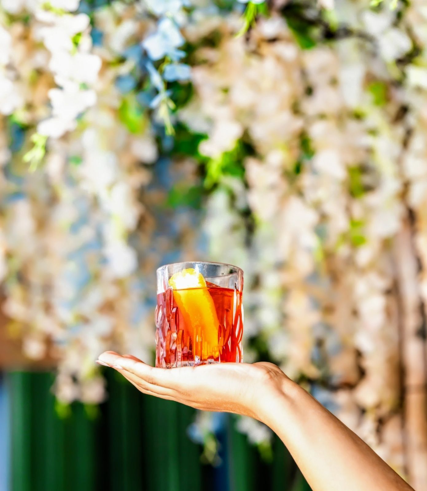 Hand holding a rocks glass with a red cocktail and orange peel against a floral background.
