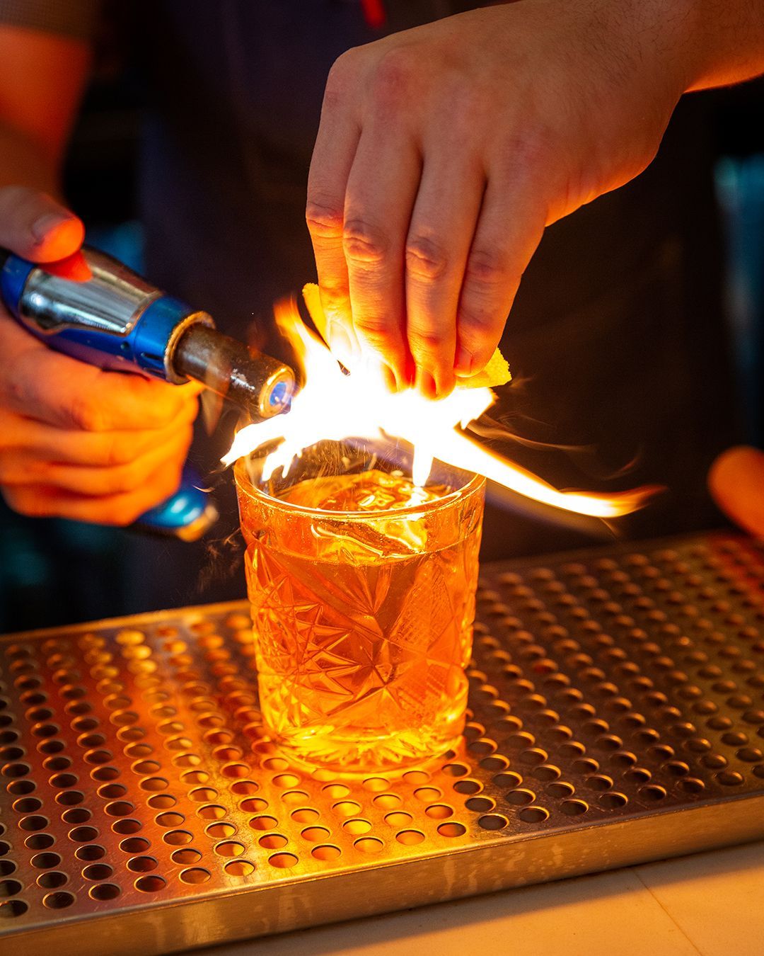 Bartender torches orange peel over cocktail glass, flames illuminating drink and tray.
