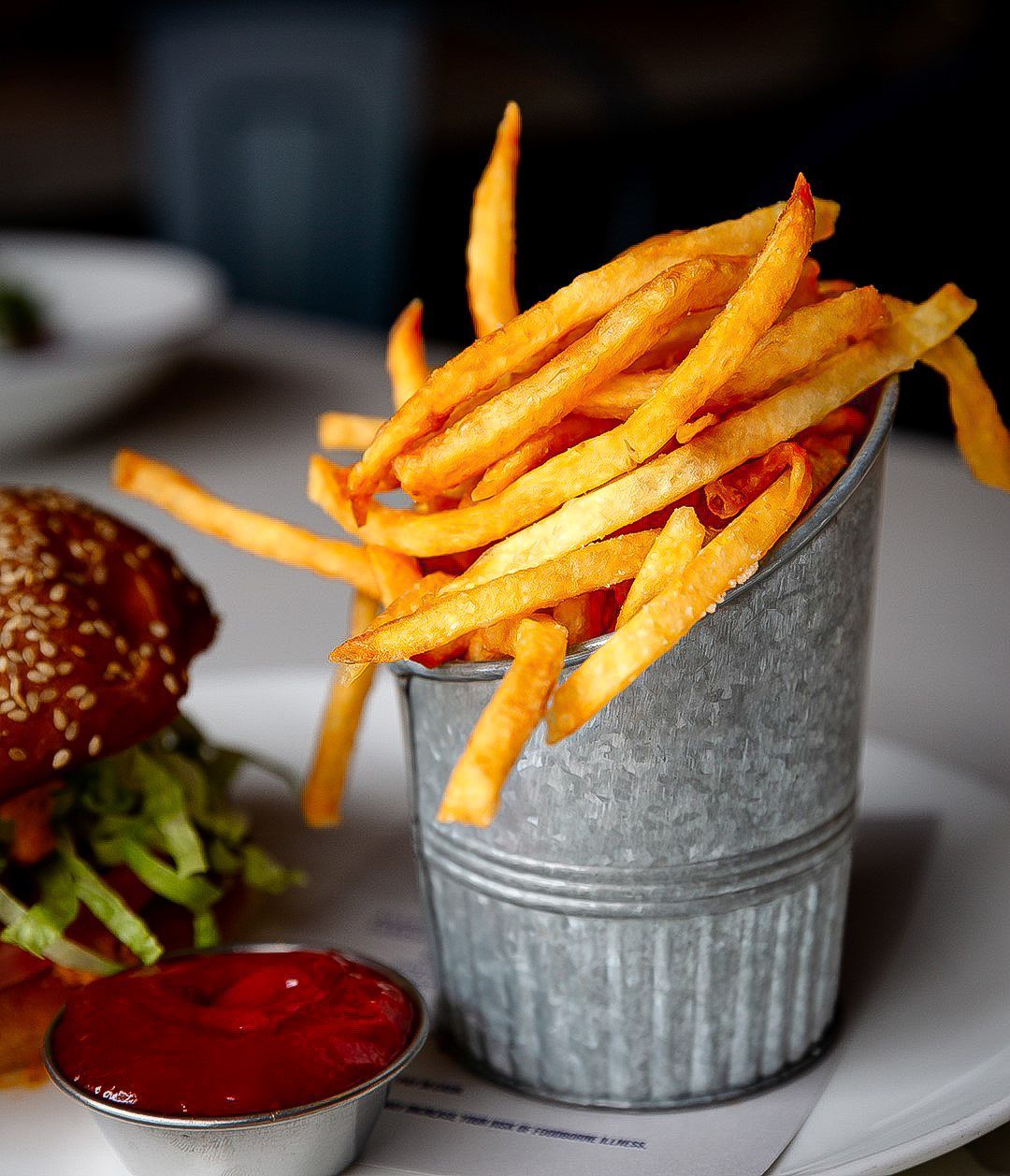 Golden french fries in metal bucket, burger with sesame bun, and ketchup on a plate.