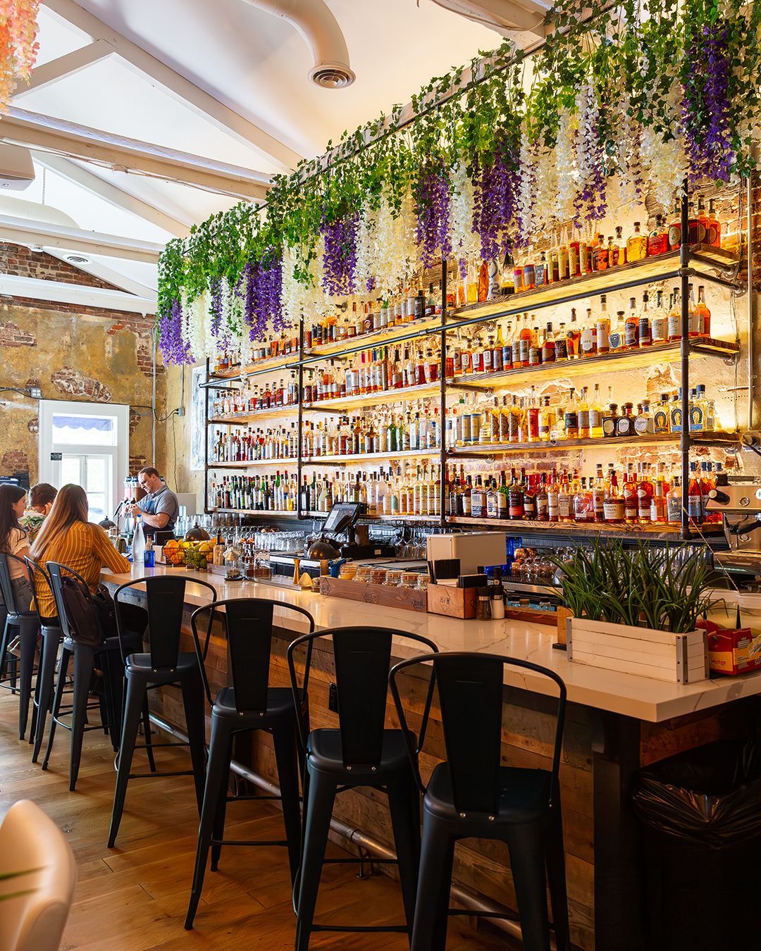 Bar interior with shelves of liquor, bar stools, and hanging floral decorations.