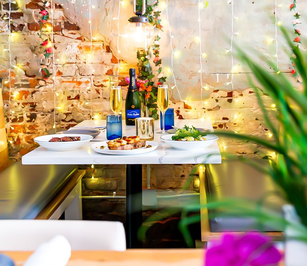 Romantic dinner setting with a table, food, drinks, and string lights against a brick wall.