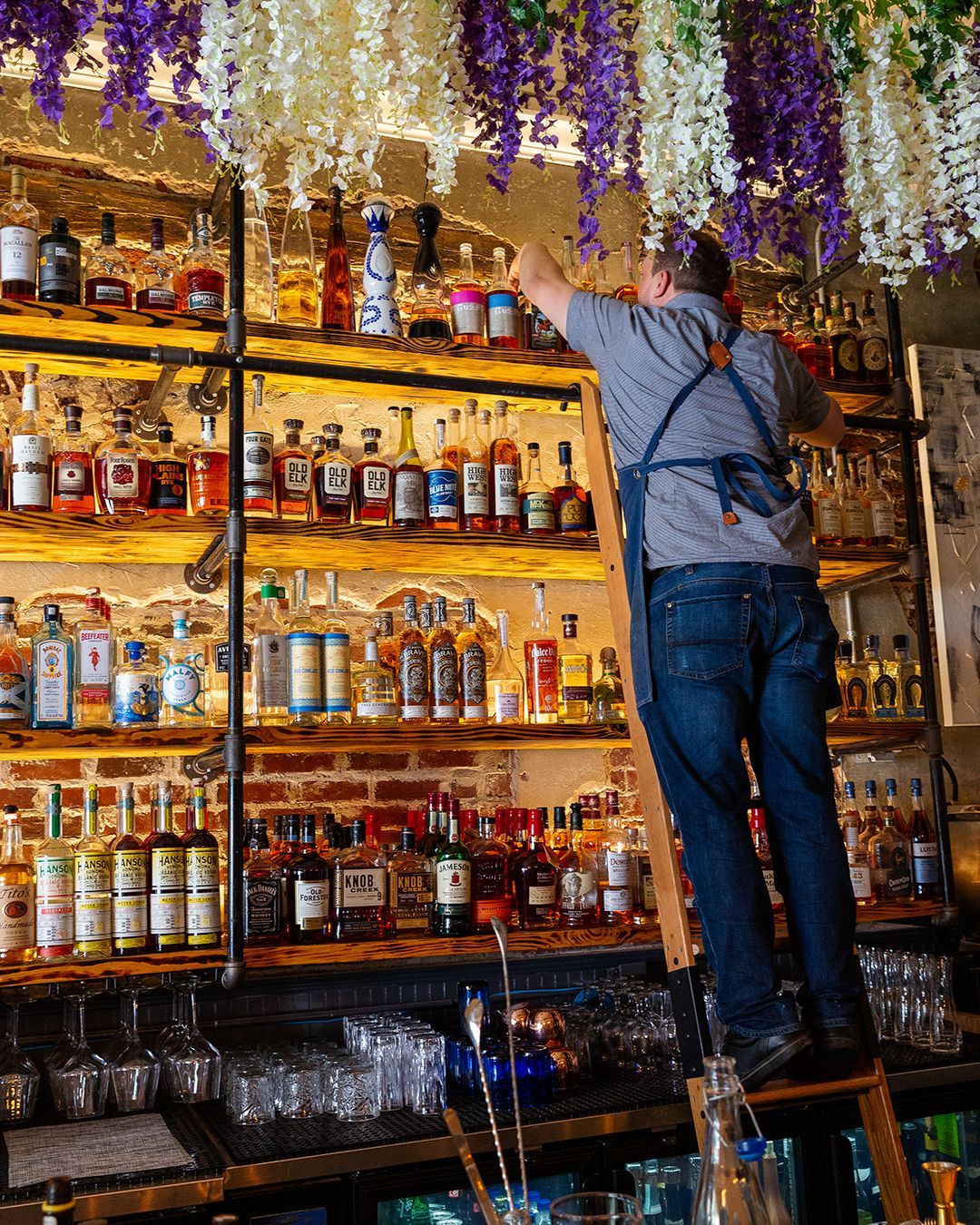 Bartender on ladder reaching for bottle on a bar shelf, surrounded by alcohol bottles and hanging purple flowers.