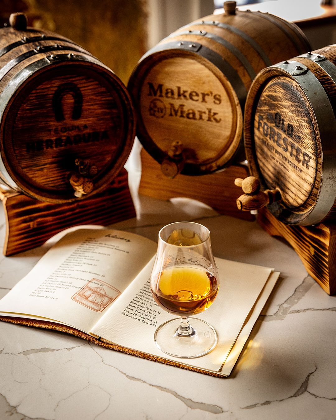 Three bourbon barrels behind a glass of amber liquid and a book on a marble surface.