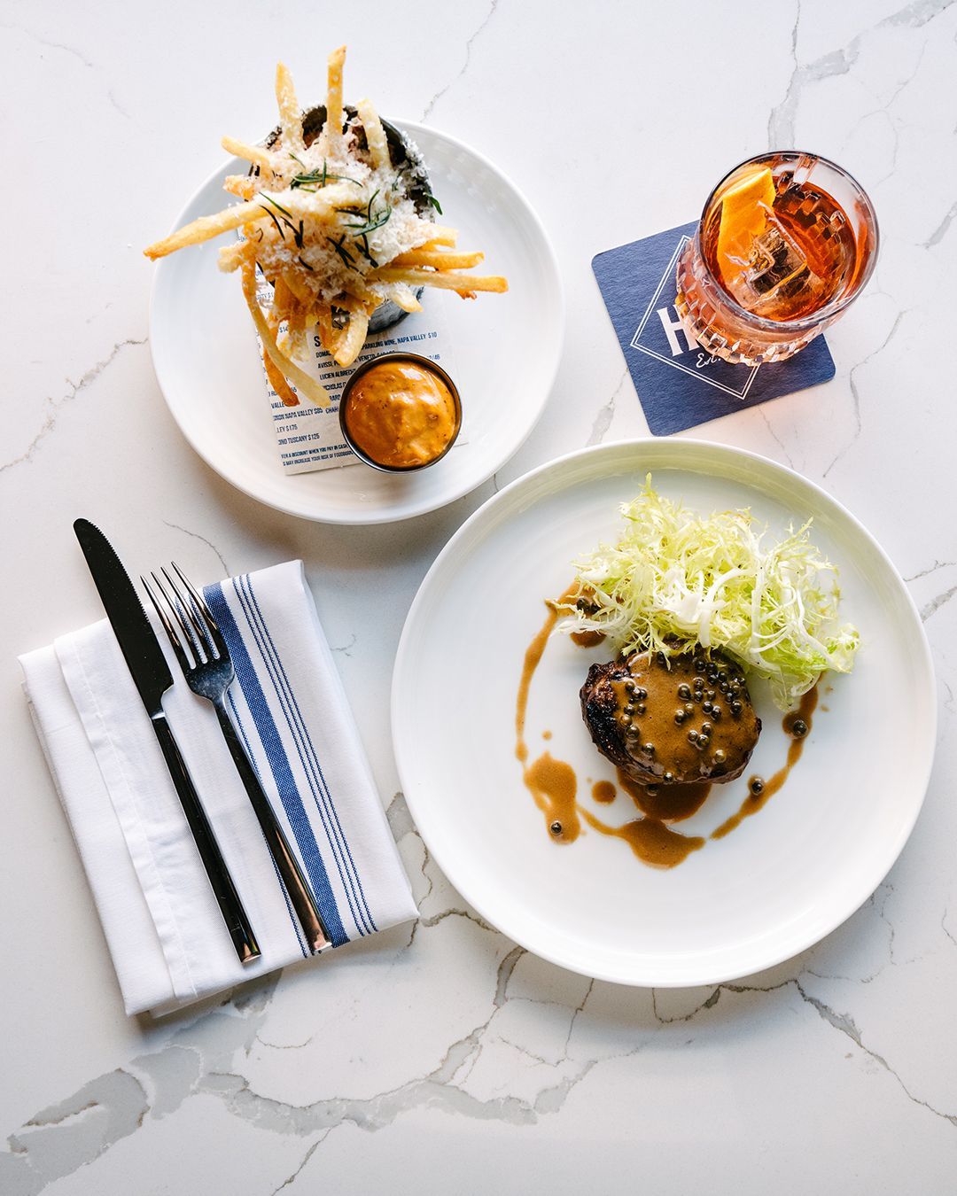 Plated dishes with fries, a burger, and a cocktail on a marble surface.