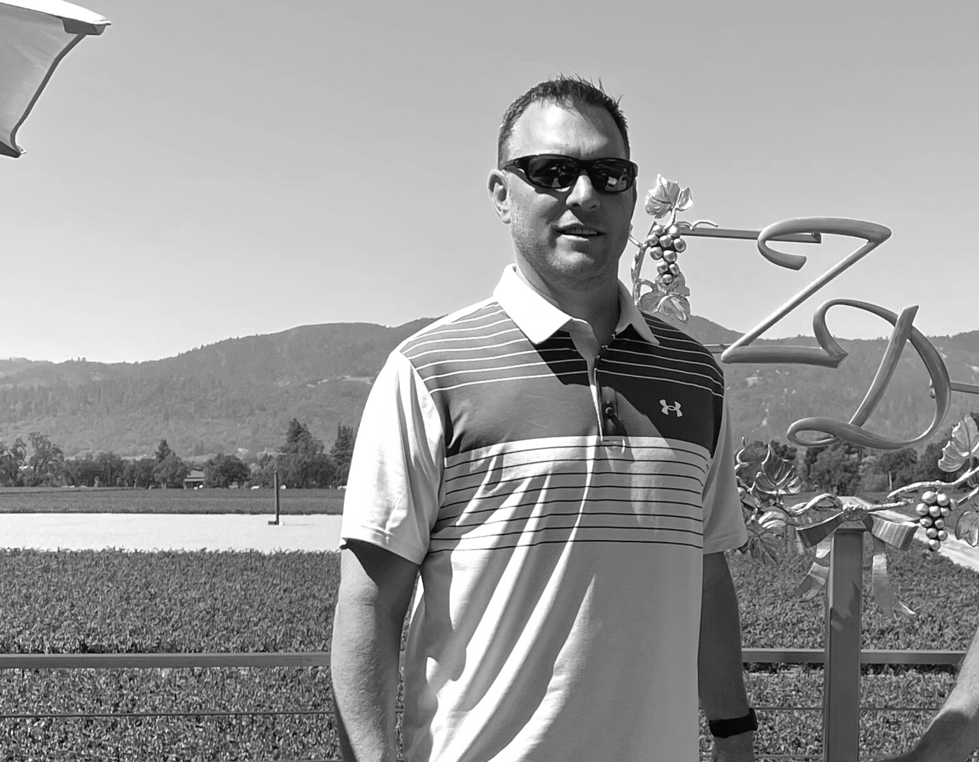 Man in sunglasses stands outside near a vineyard.