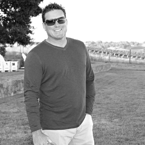 Man in sunglasses smiles outside, hands in pockets. V-neck shirt, grassy area, fence in background.