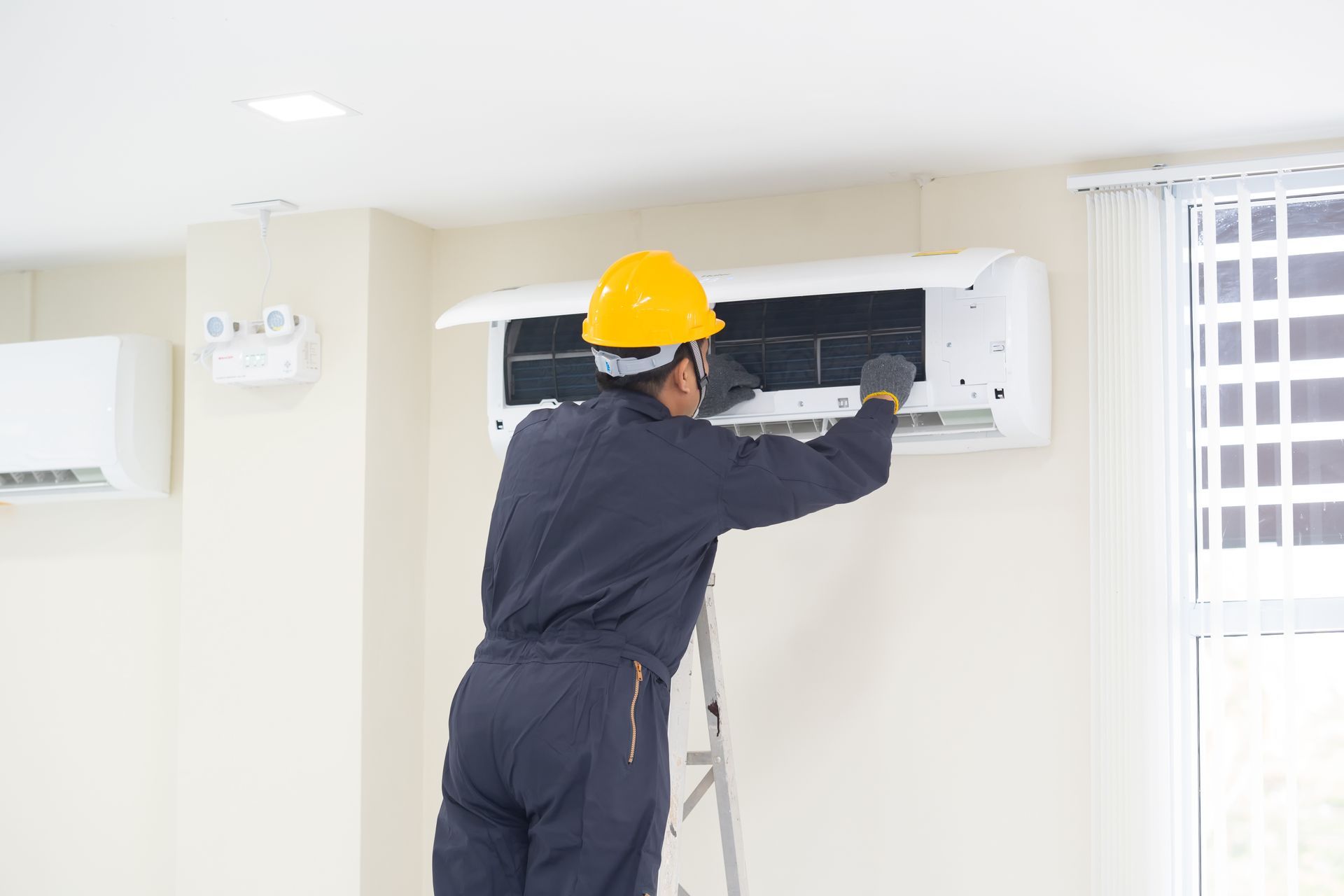 Technician in hard hat servicing a wall-mounted air conditioner indoors.