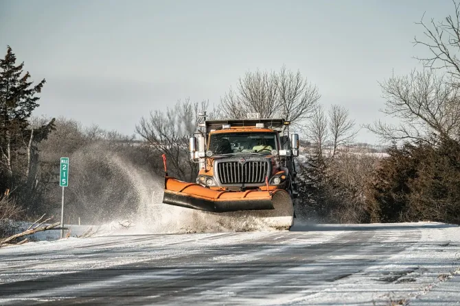 Orange snowplow clearing a snowy road, spraying snow under a gray winter sky