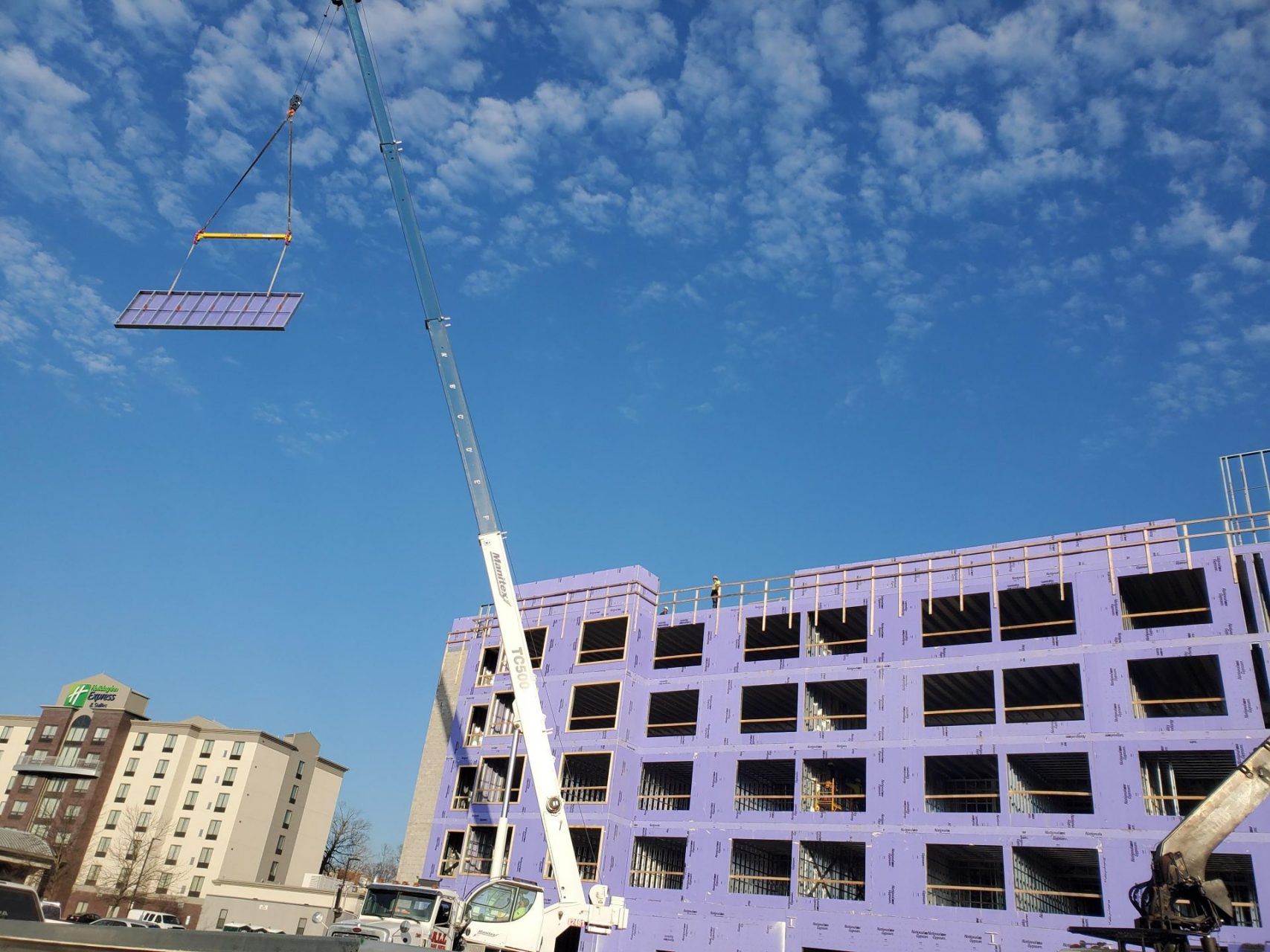 A crane is lifting a large steel section of the  building high in the air in front of a large building under construction.
