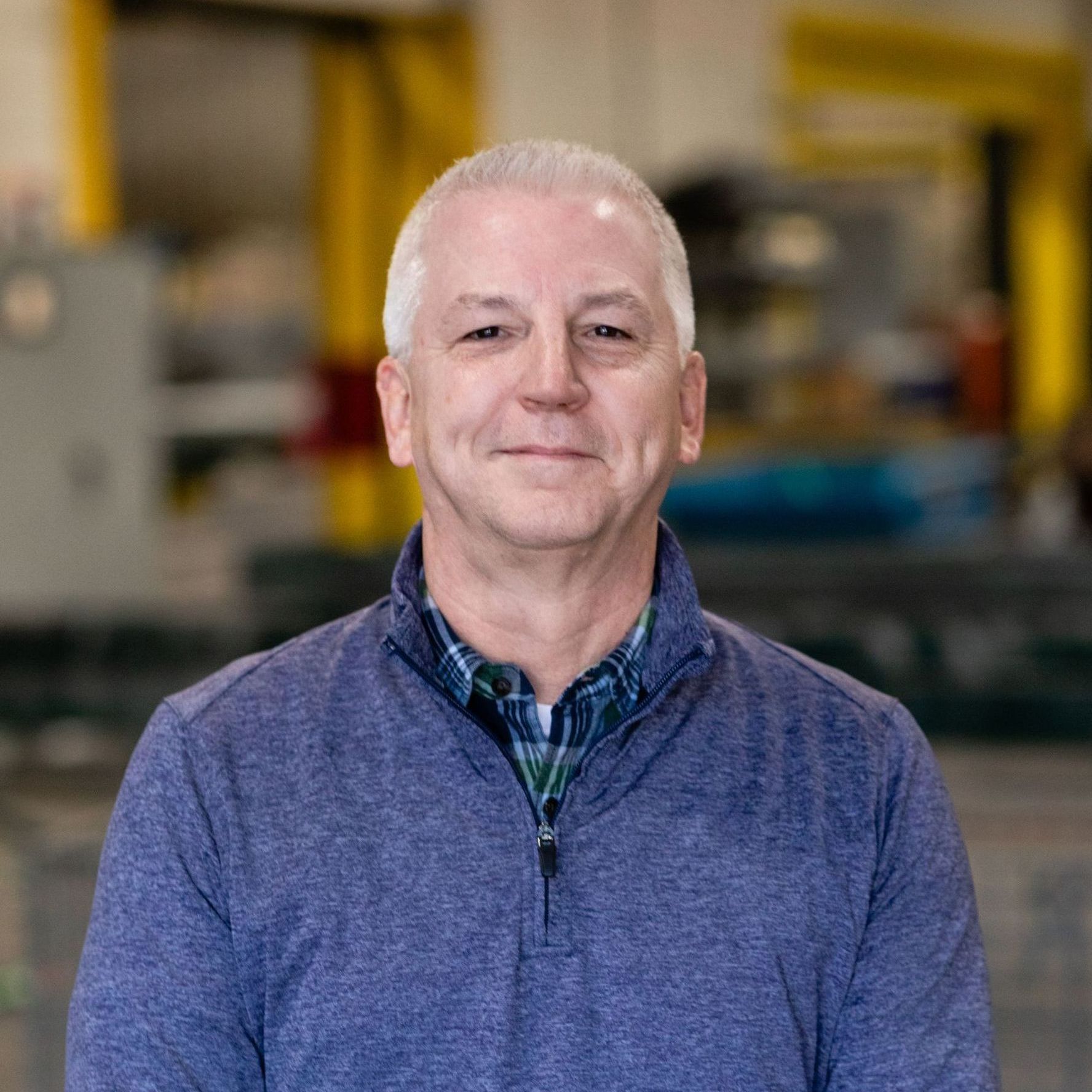 A man wearing a blue sweater and a plaid shirt is smiling.