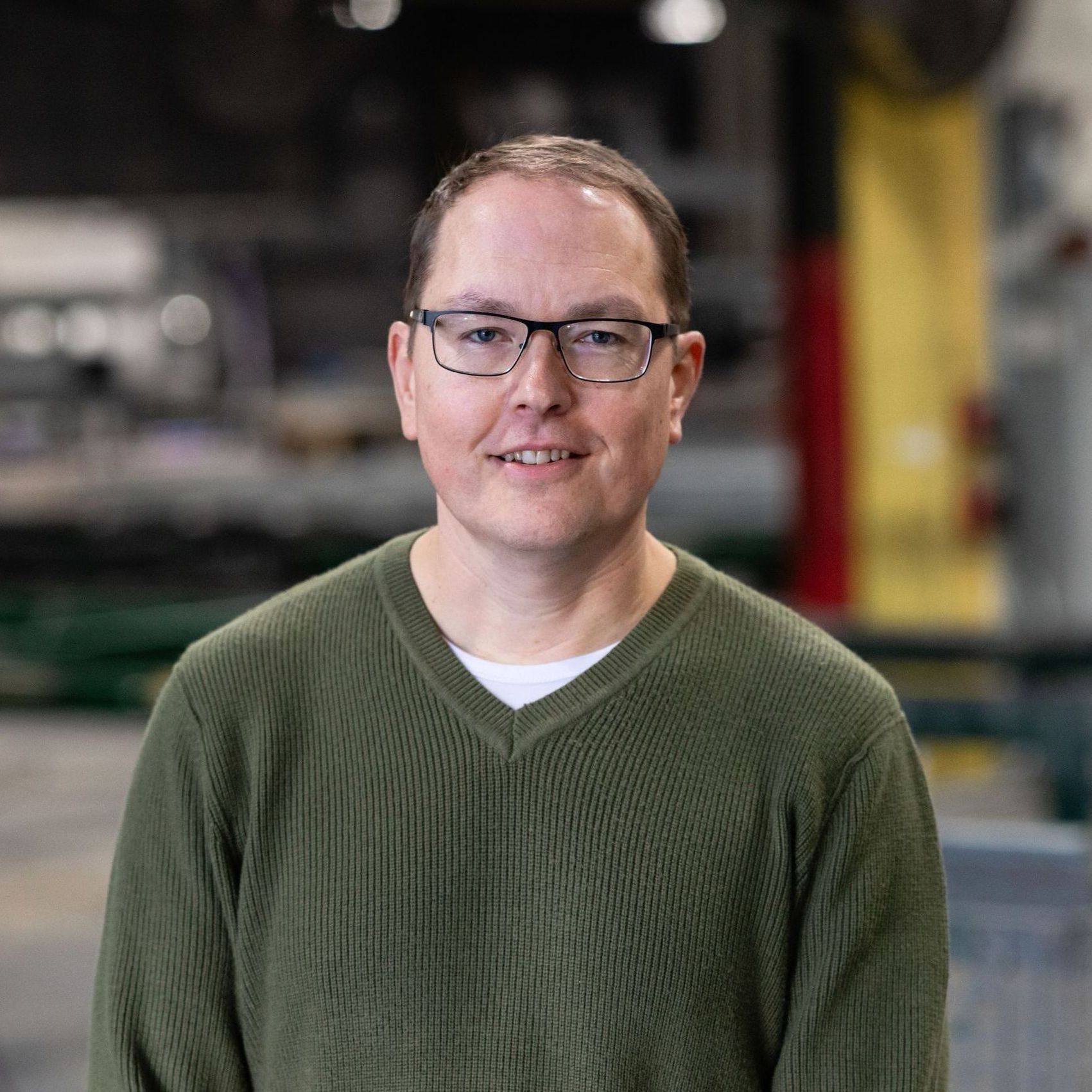 A man wearing glasses and a green sweater is smiling and standing in a factory.