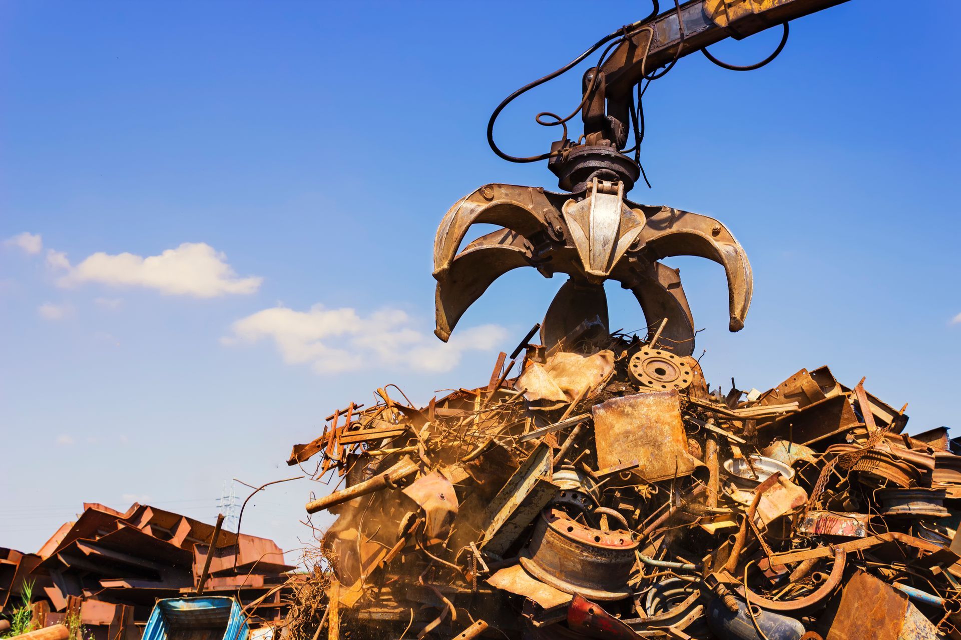 Industrial claw lifting scrap metal at a junkyard under a clear blue sky.