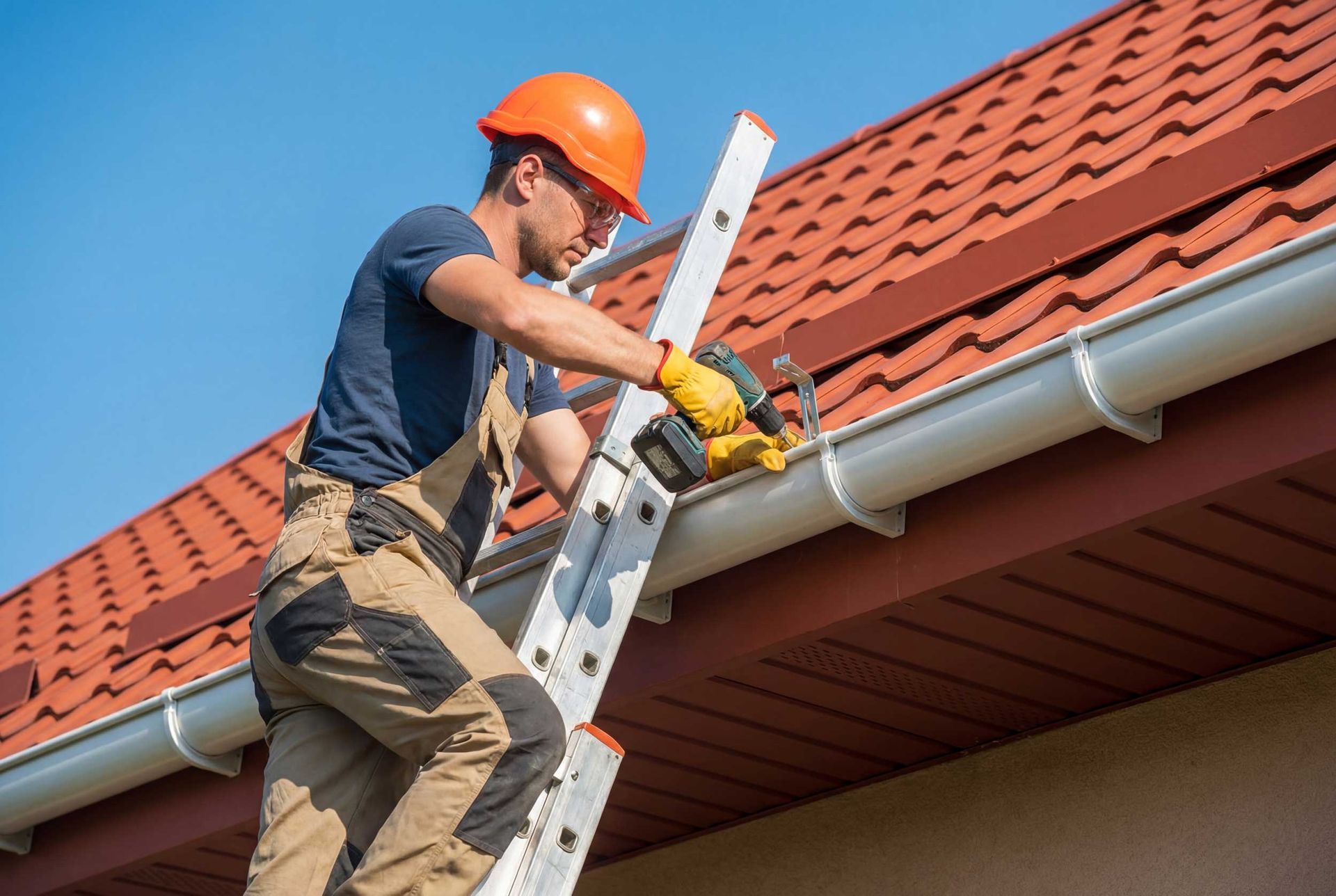 An expert gutter repair contractor on a ladder fixing a white residential gutter with a power drill.