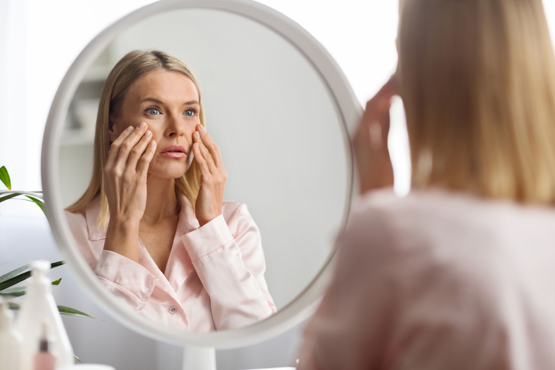 Woman looking in mirror, touching face, concerned expression. Pink robe, white frame.