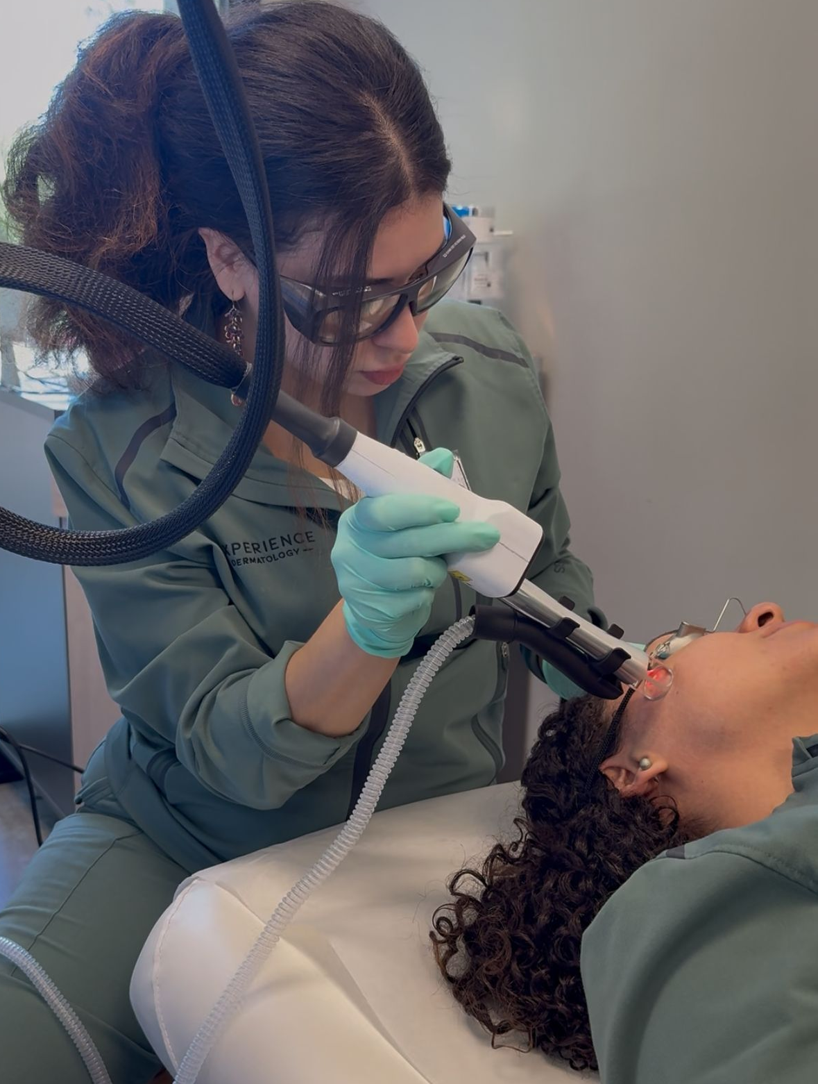Person in teal scrubs using a medical device on a person lying down, both wearing eye protection.