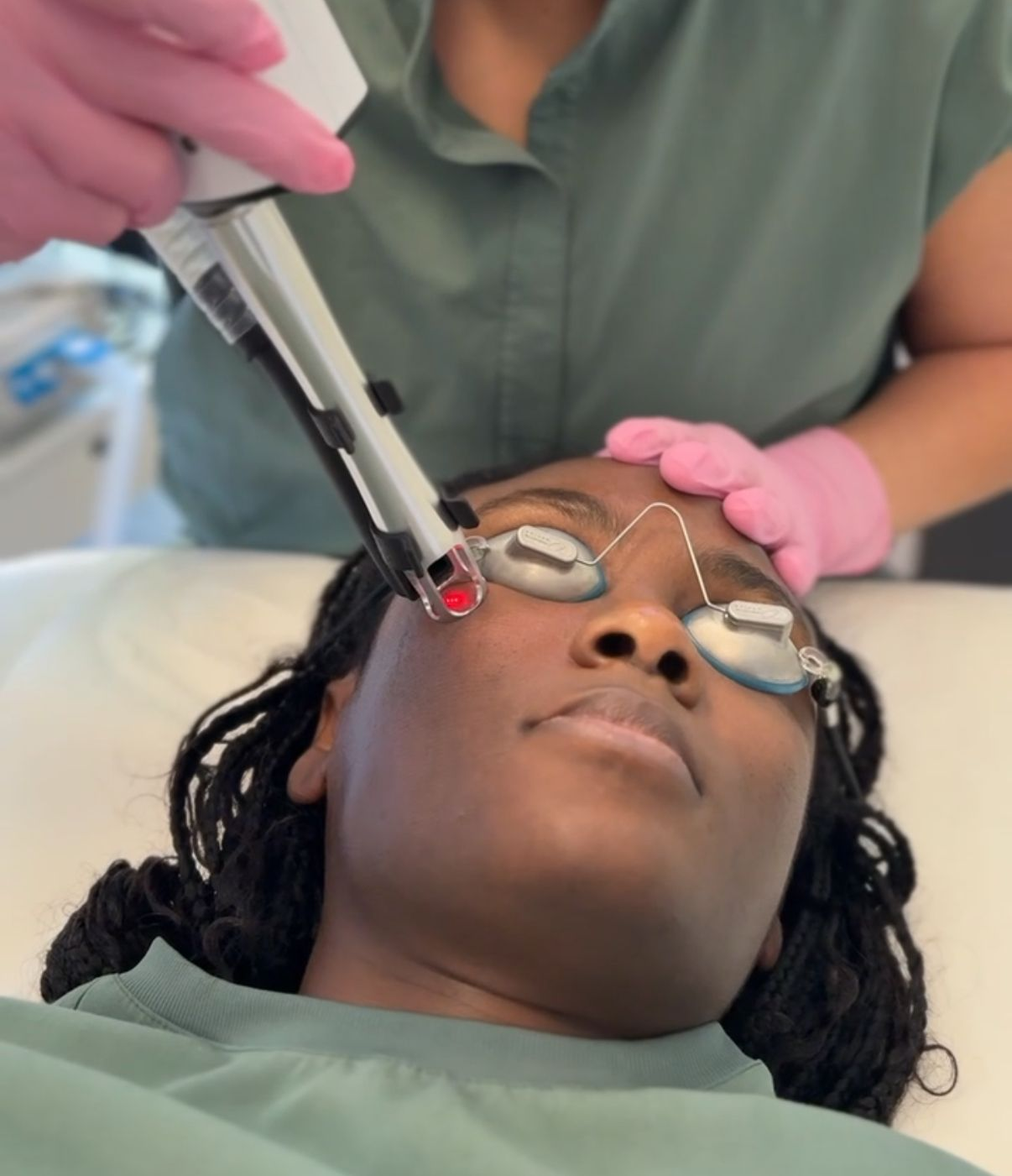 Woman in medical attire using a laser device on a patient's face in a clinical setting.