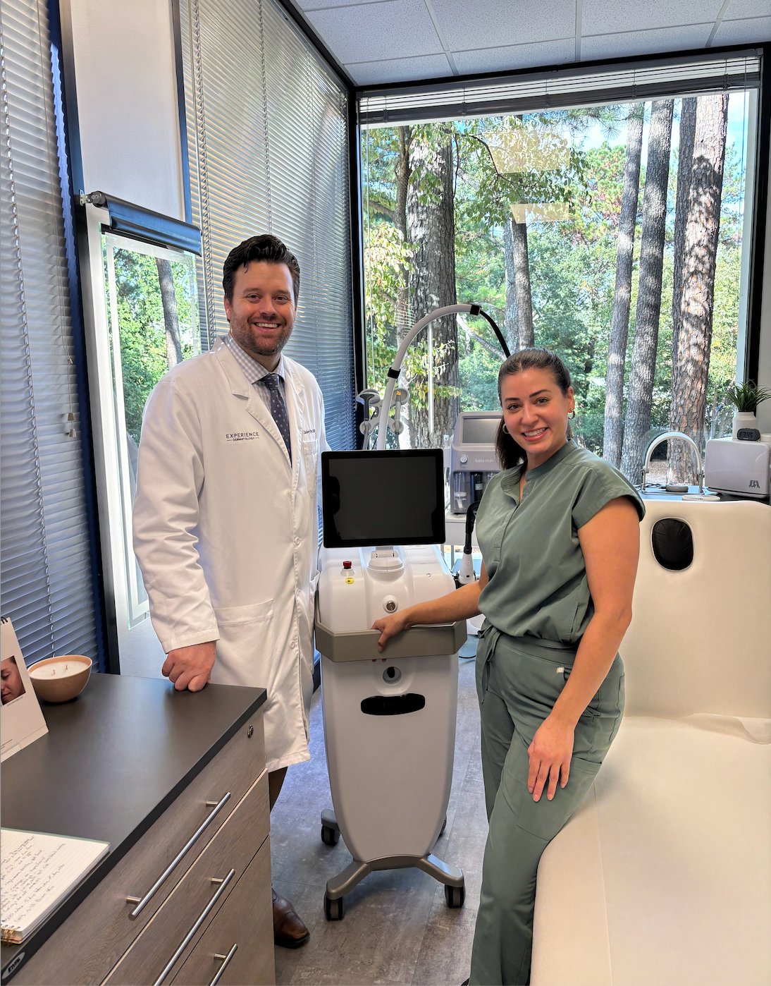 A doctor and assistant stand near a medical device in a clinical setting. Both smile.