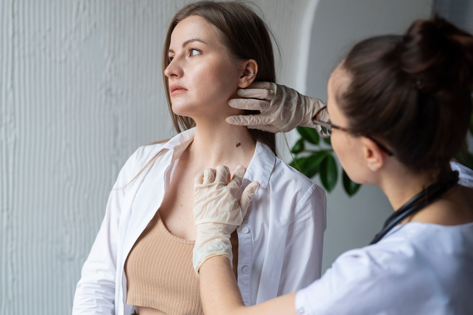 A doctor examines a woman's neck for moles. Light-skinned woman, both wearing white and beige.