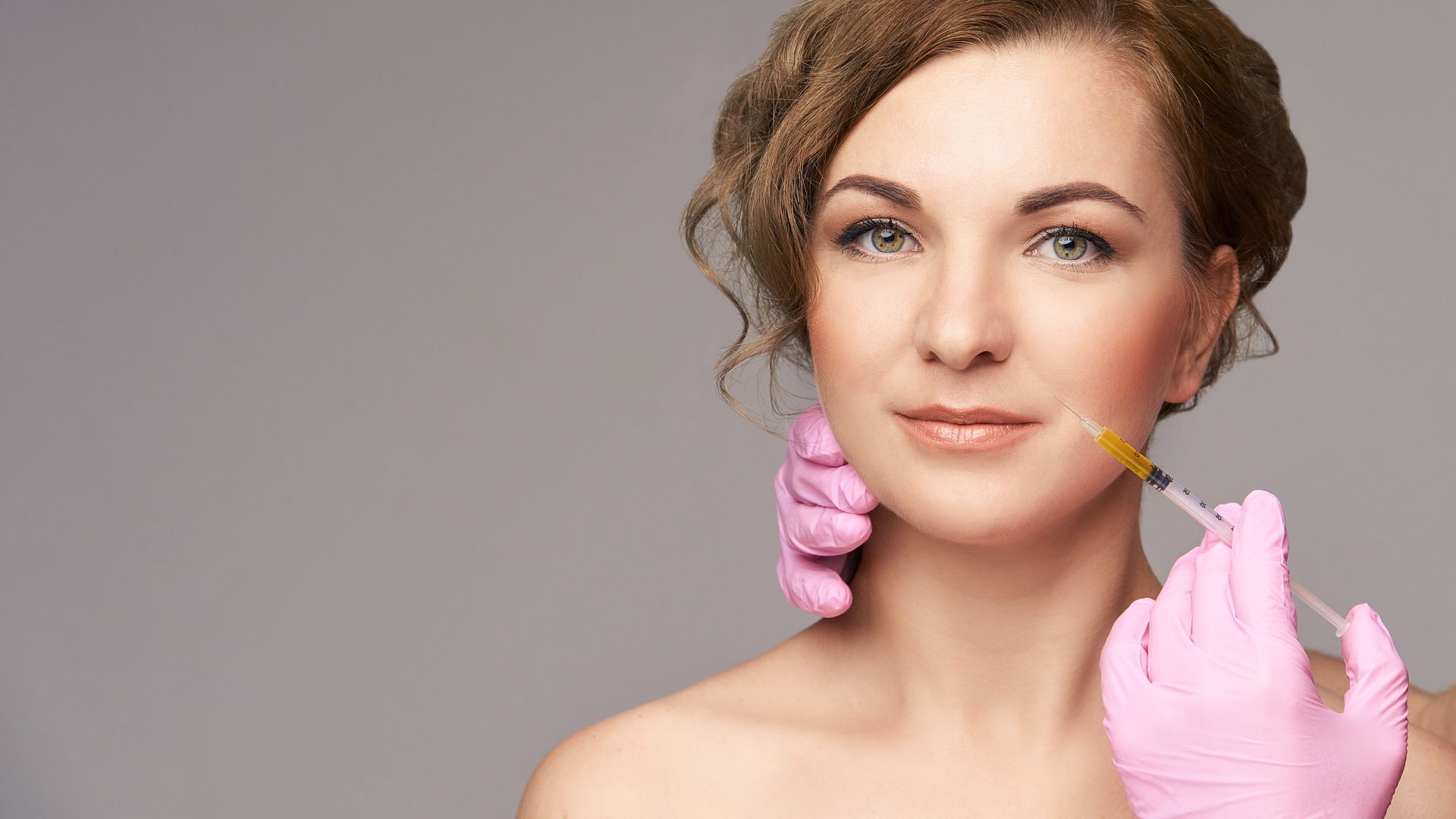 Woman receiving a facial injection, wearing pink gloves, in front of a gray background.