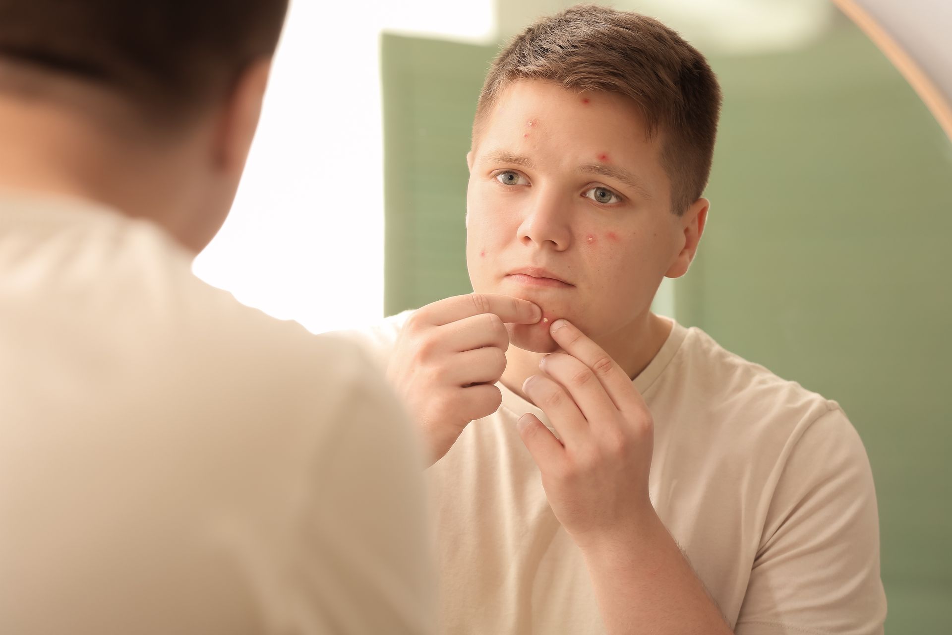 A person looking in a mirror, squeezing a pimple on their chin in a bathroom.