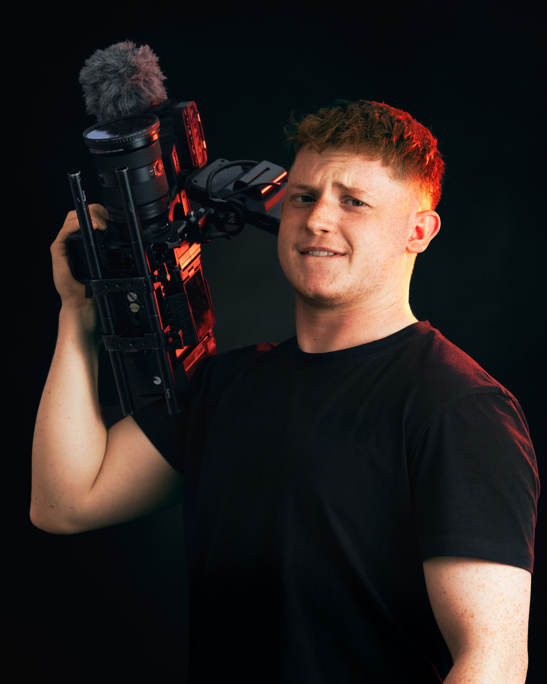 Man with red hair holding a camera, looking confused, with a dark background.