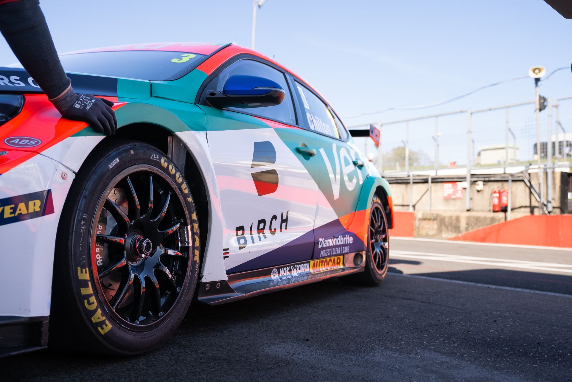 Team Vertu race car on a track, low angle view with visible sponsors and a blue sky background