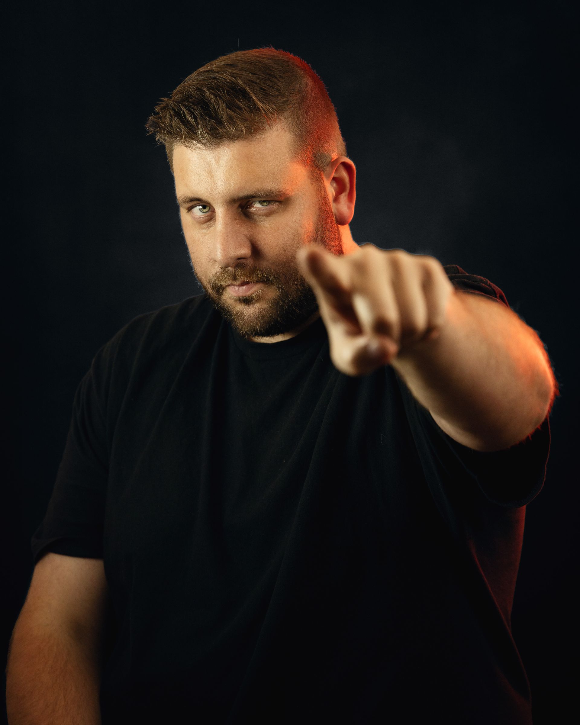 Man with beard, in black shirt, points directly at viewer with a serious expression against a dark background.