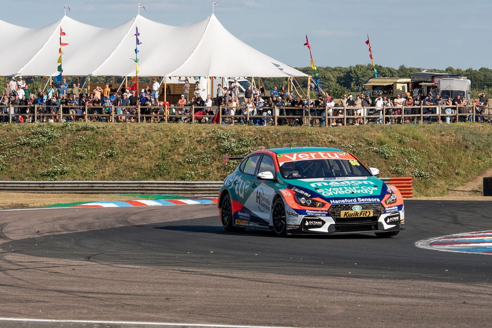 Colorful EXCELR8 race car taking a turn on a track with tents and spectators in the background