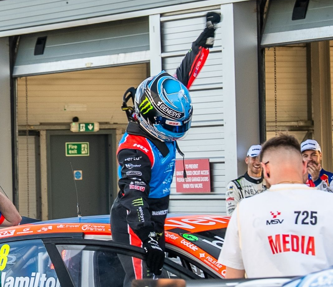 Nick Hamilton in blue helmet raises arm beside orange car in pit lane, with media crew nearby
