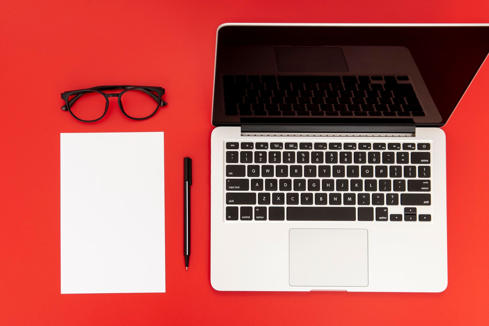 A laptop , notebook , pen , glasses and a notepad on a red background.