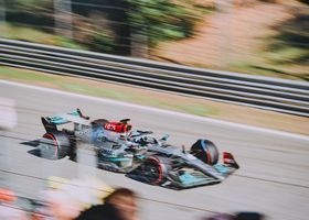 Two Formula 1 cars racing side by side on a blurred track, one silver and teal, one dark blue and red.