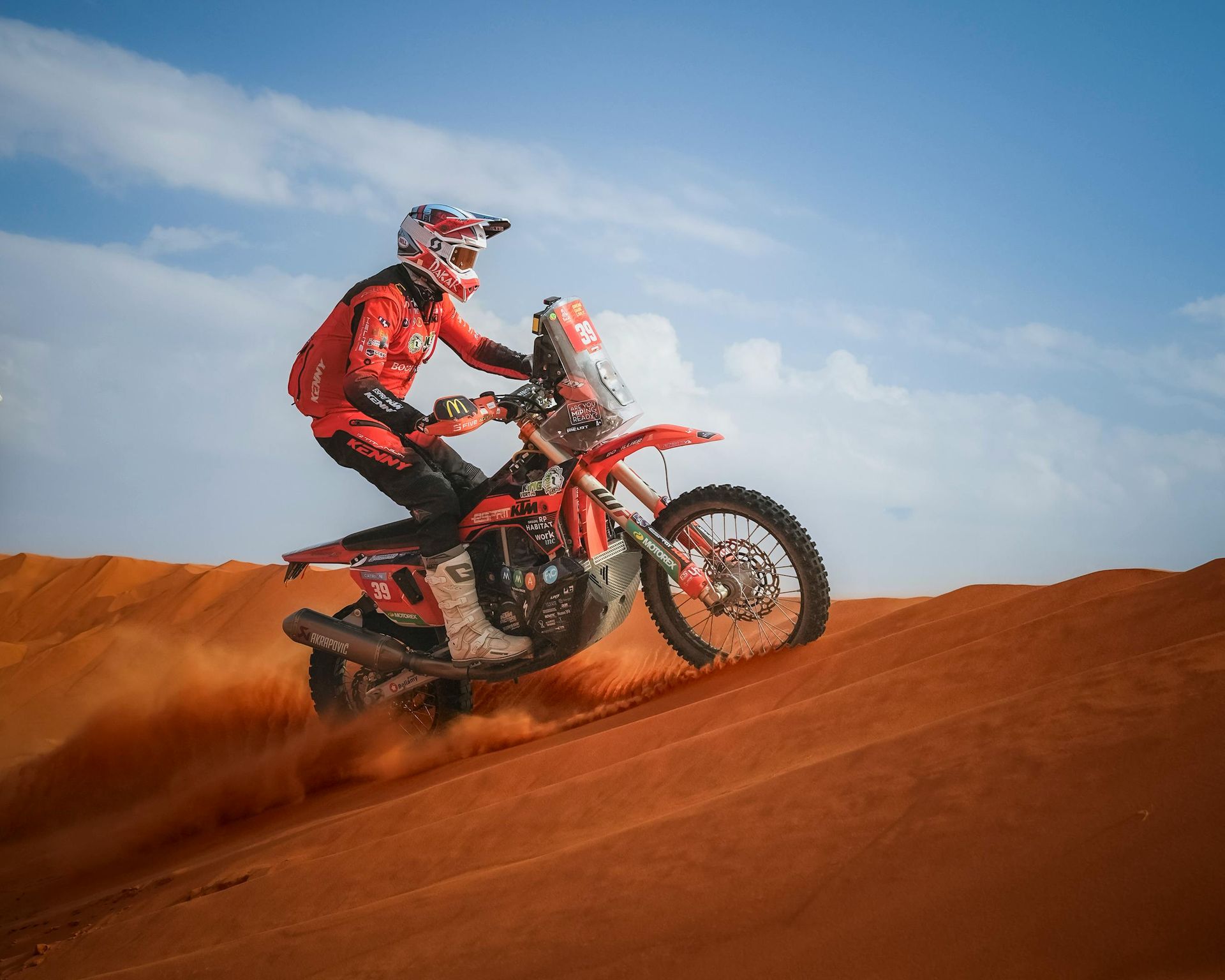 A motorcyclist in red riding gear speeds across sand dunes under a blue sky, kicking up dust behind the bike.