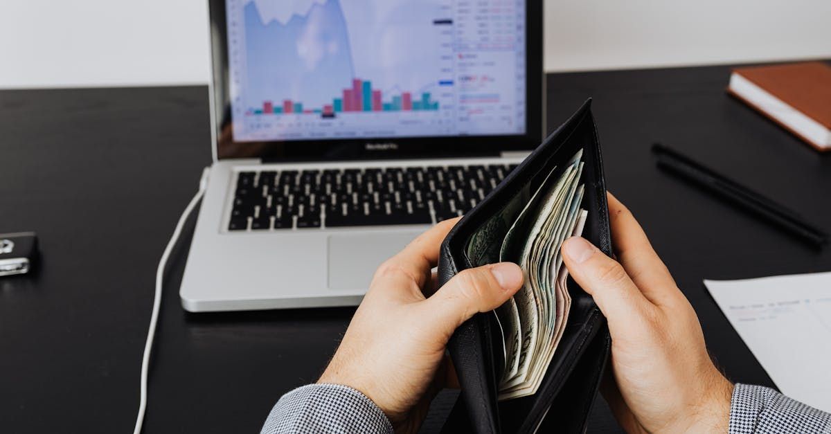 Person holding open wallet filled with cash, laptop and documents on desk in background.