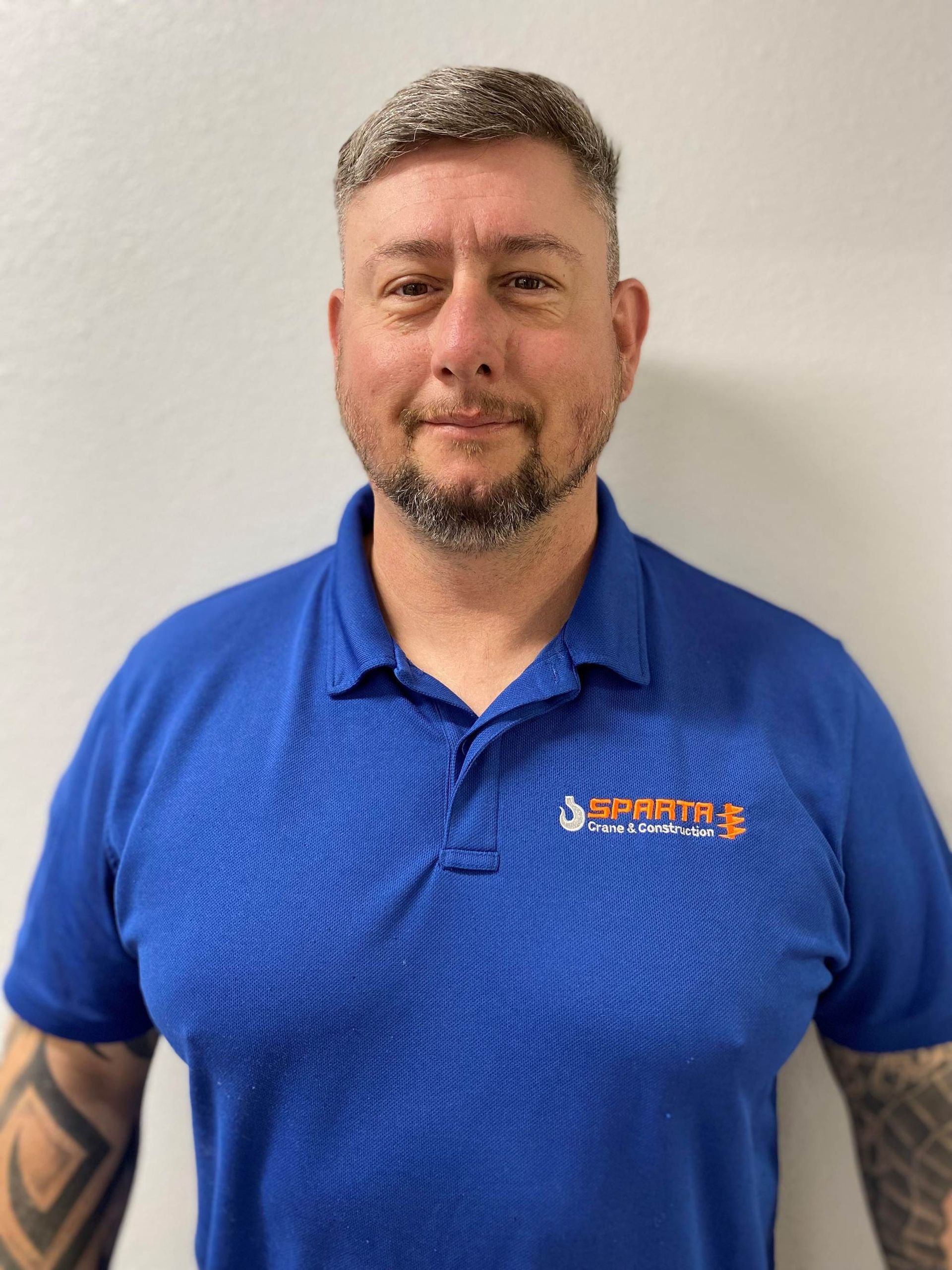 A man with a beard and tattoos is wearing a blue polo shirt.
