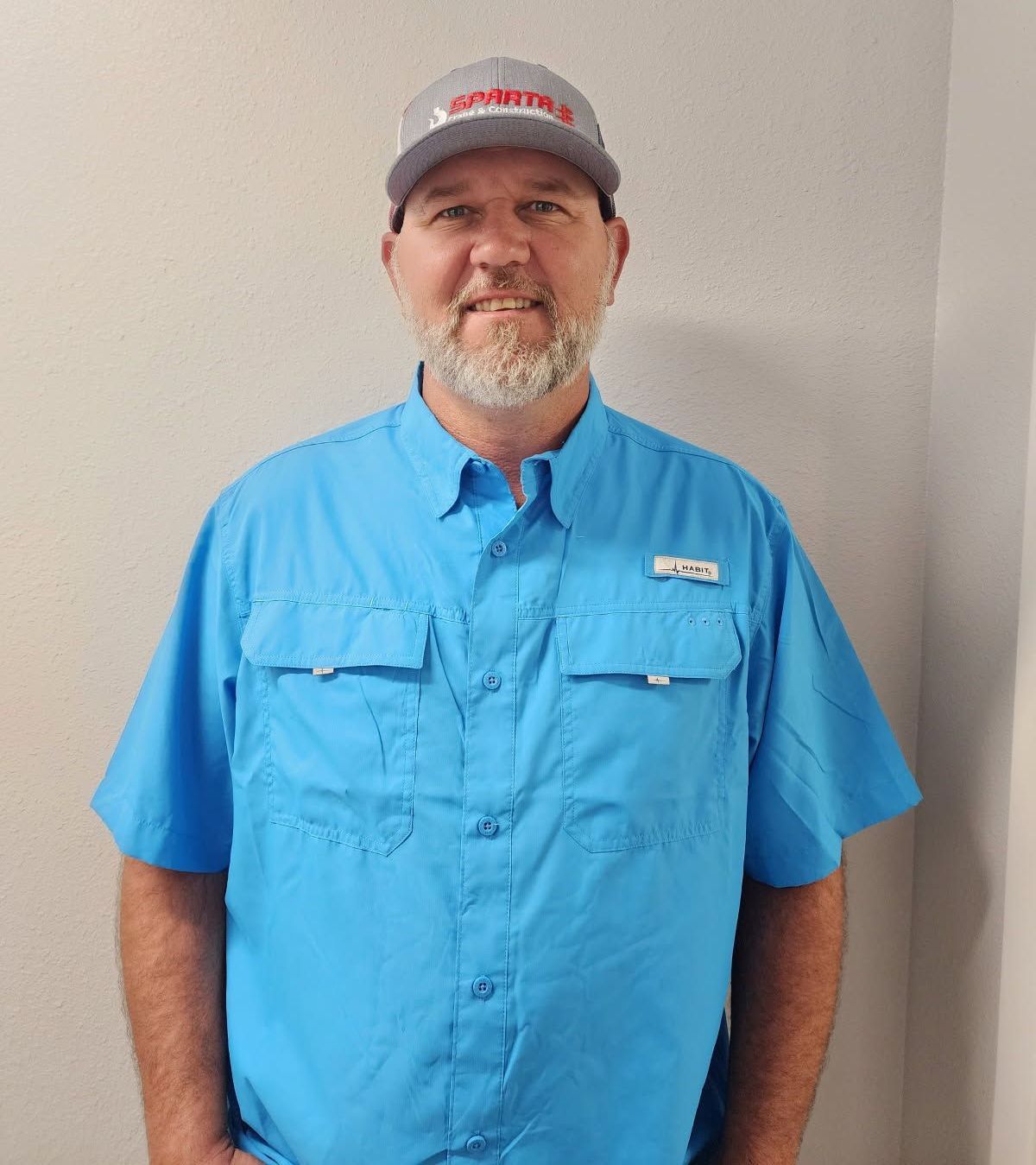 A man wearing a blue shirt and a hat is standing in front of a wall.