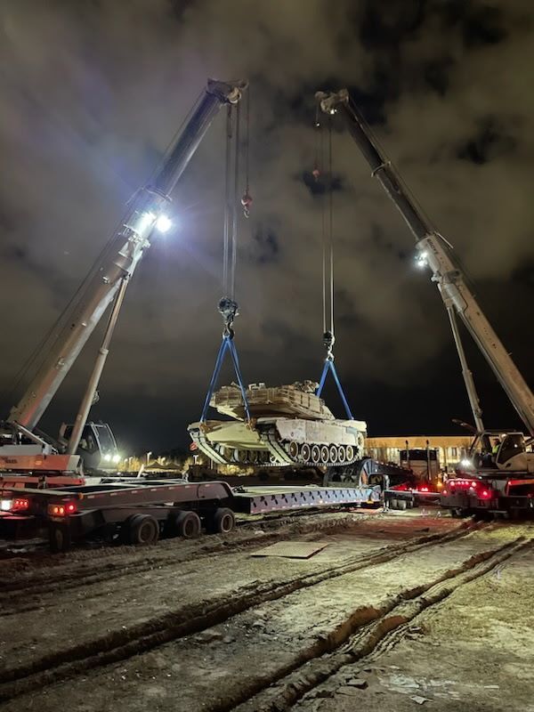 A tank is being lifted by two cranes at night.