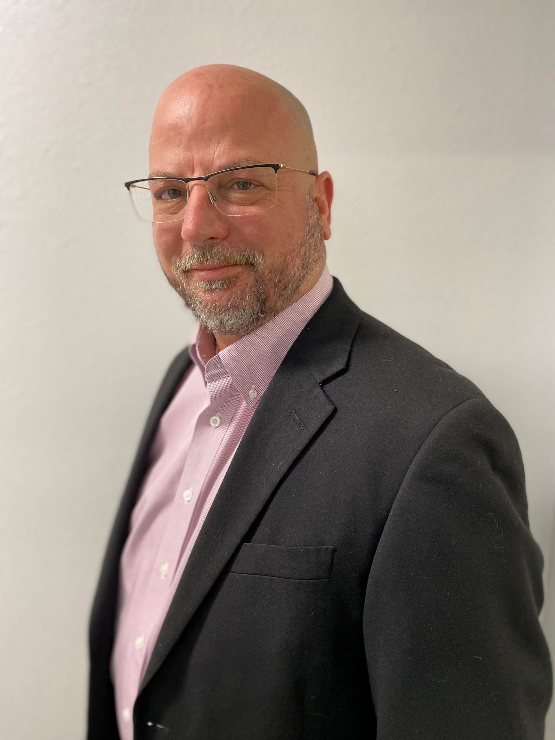 A bald man wearing glasses and a suit is standing in front of a white wall.